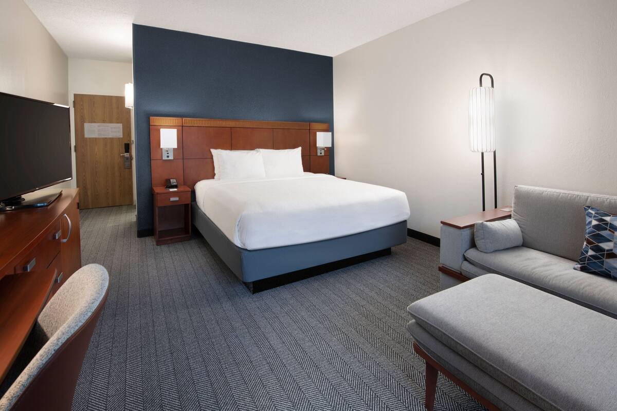 A hotel room with a large bed, a desk with a flat-screen TV, a sofa, and a standing lamp.
