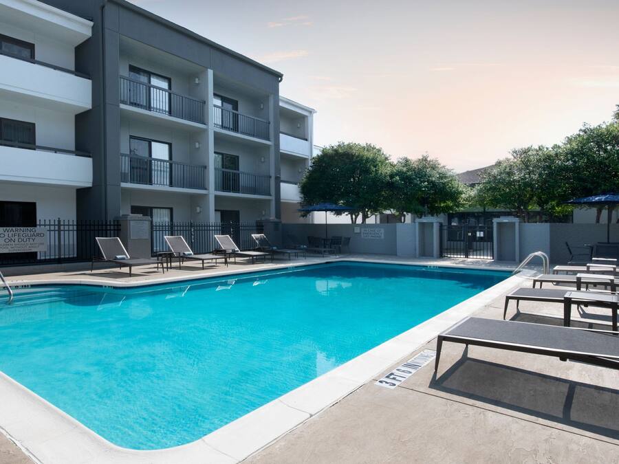 Una piscina con sillones de descanso y un edificio de varios pisos con balcones.