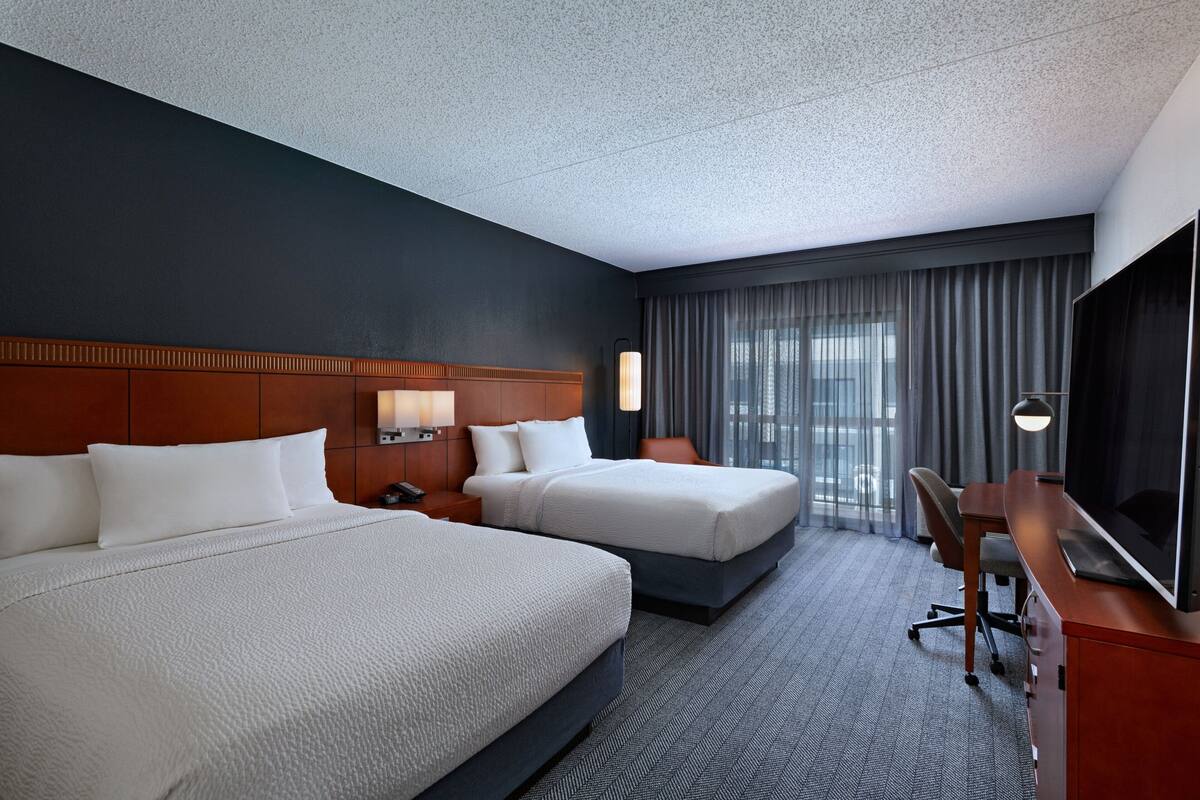 A hotel room with two beds, a desk, a TV, and a large window with curtains.