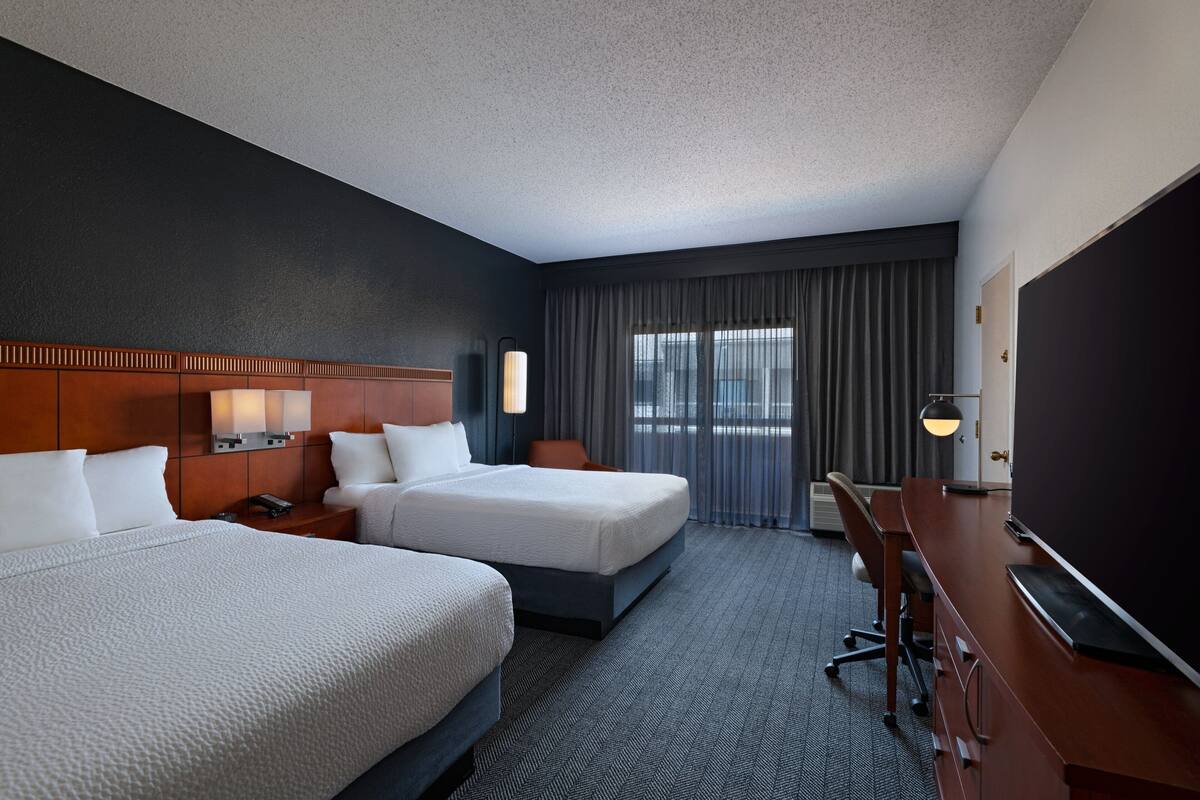 A hotel room with two beds, a desk, a TV, and a window with curtains.
