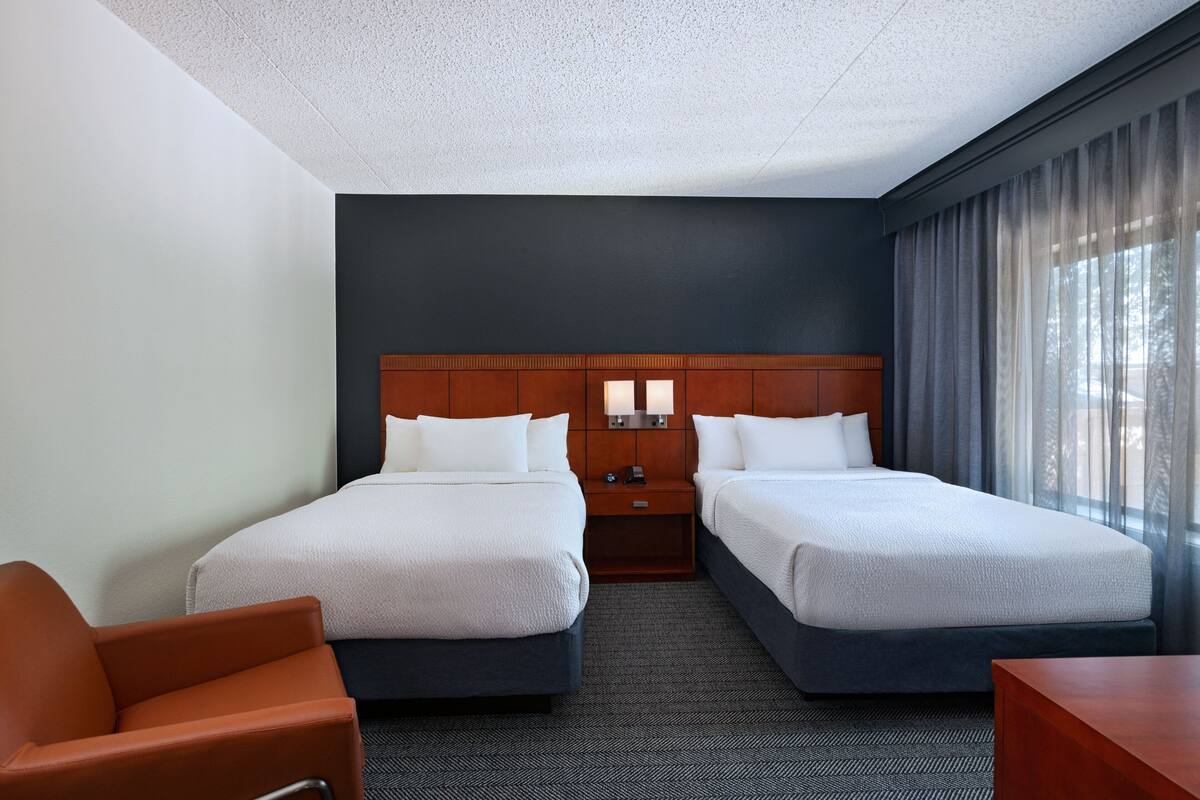 A hotel room with two beds, a wooden headboard, a nightstand with a lamp, a desk, and a chair.