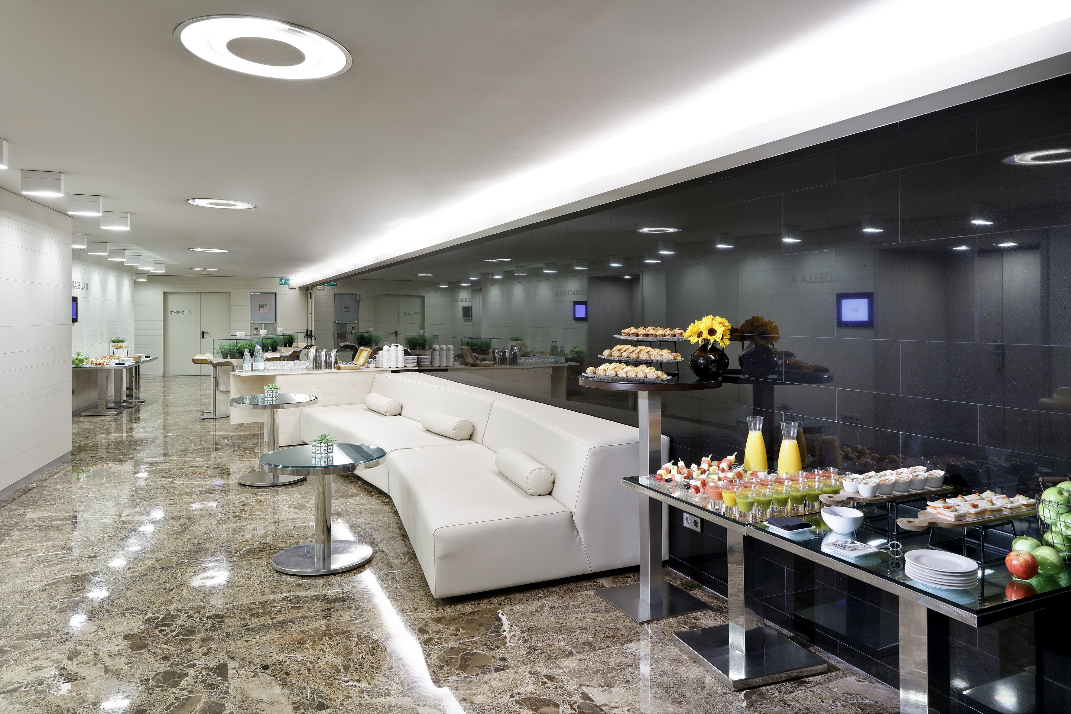 Un vestíbulo de hotel moderno con suelo de mármol, un sofá blanco y una zona de bufé con diversas opciones de comida.