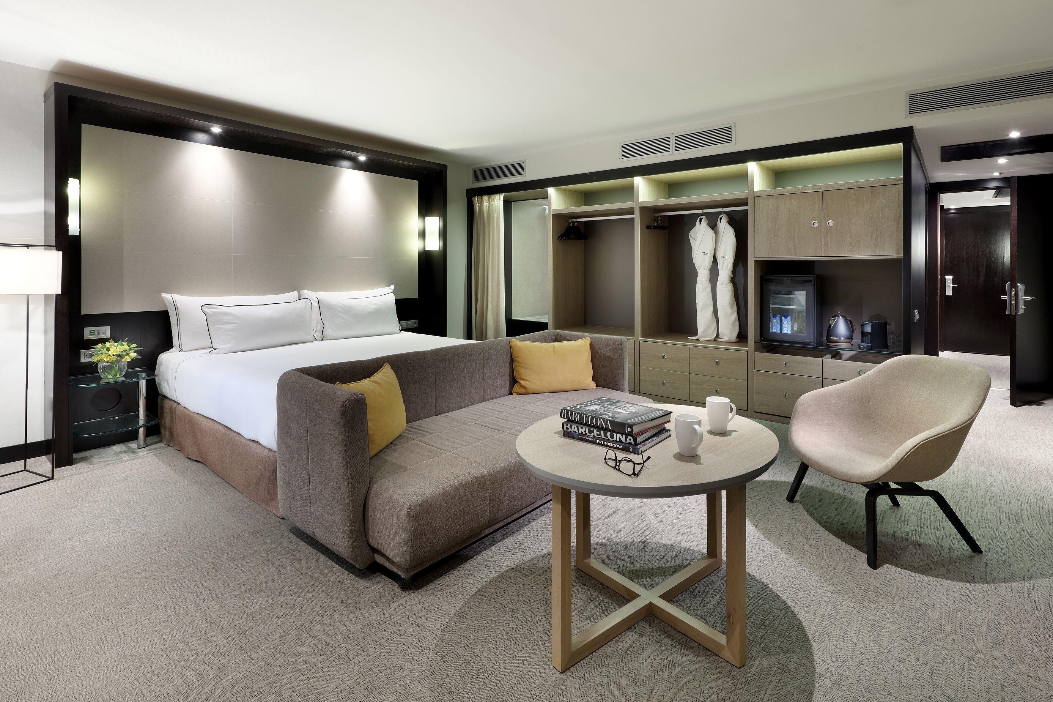 A modern hotel room with a bed, a sofa, a small table, a chair, a wardrobe, and a mirror.
