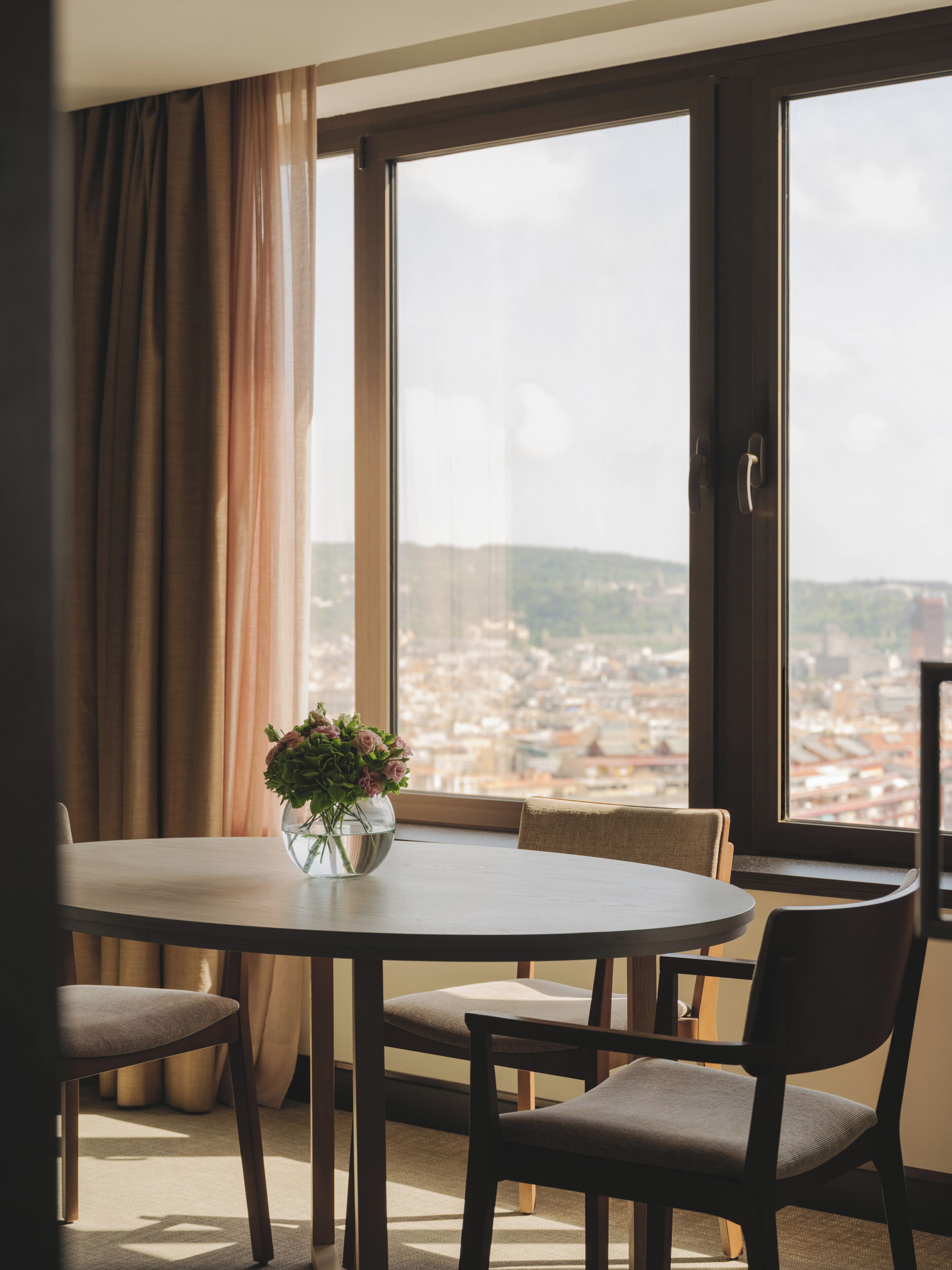 A dining area with a table and chairs, a vase with flowers, and a view of a cityscape through large windows.