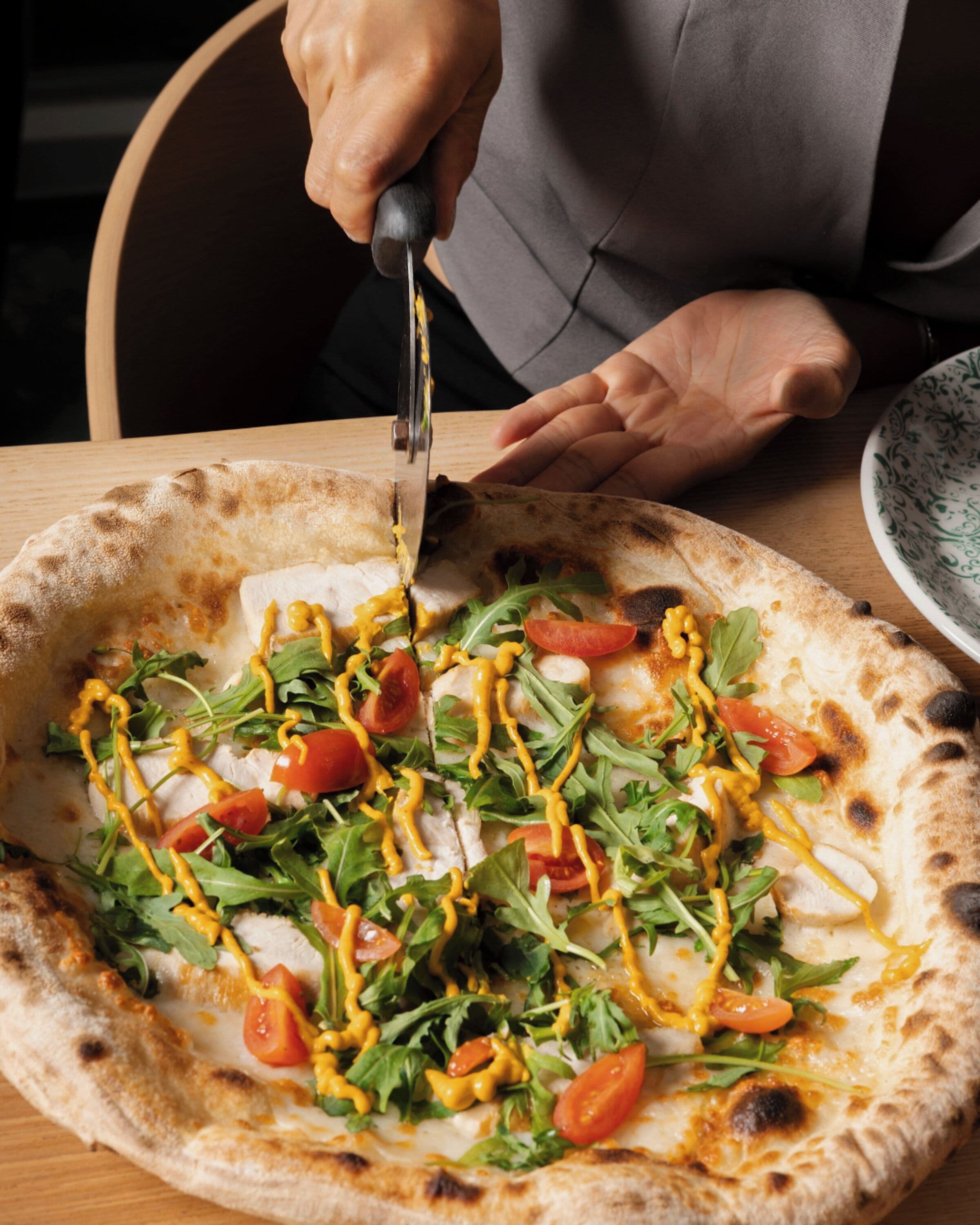 A person cutting a pizza topped with greens, tomatoes, and cheese.