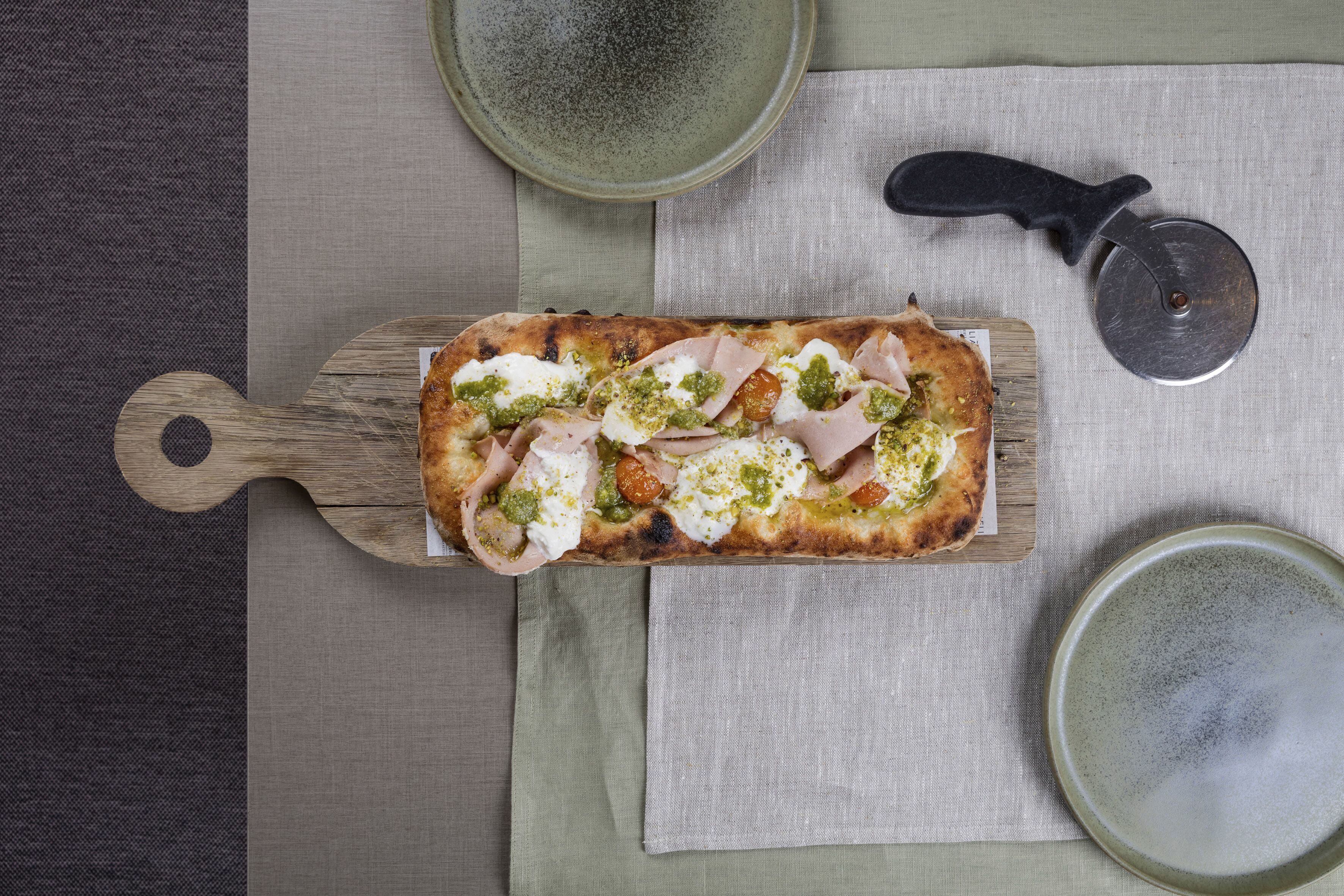 Una pizza rectangular sobre una tabla de madera, un cortador de pizza y dos platos de cerámica sobre un mantel.