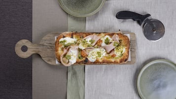 Una pizza rectangular sobre una tabla de madera, un cortador de pizza y dos platos de cerámica sobre un mantel.