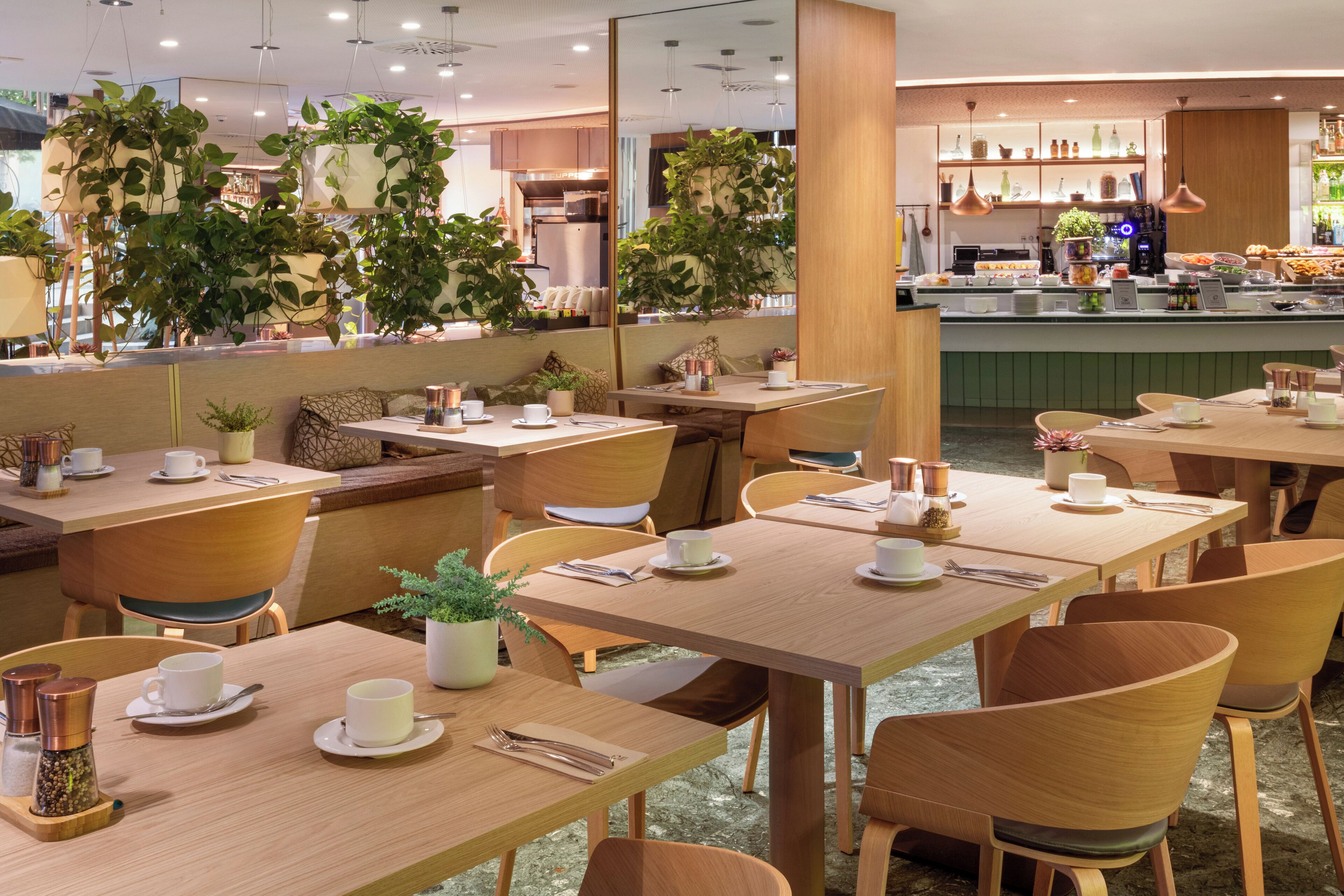 A restaurant with wooden tables and chairs, potted plants, and a buffet area in the background.