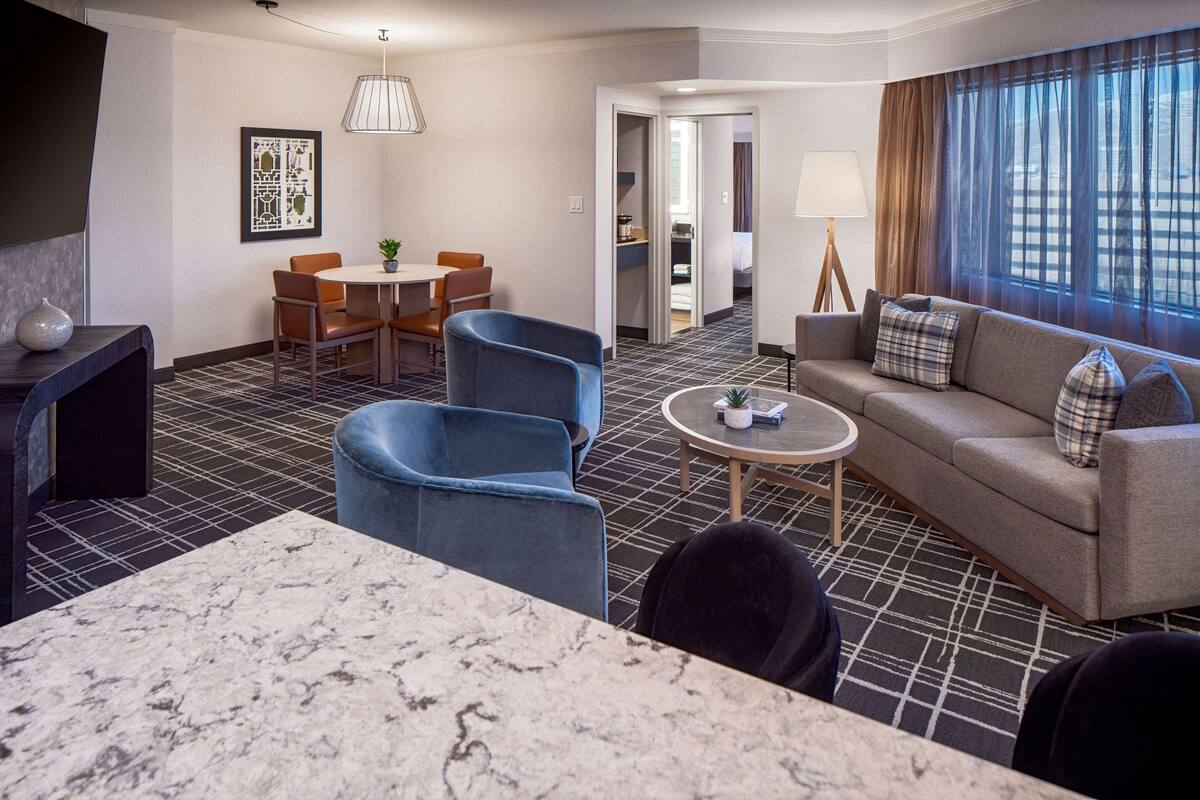 A hotel room with a marble-tiled dining area, a sofa with plaid cushions, and a view through large windows.