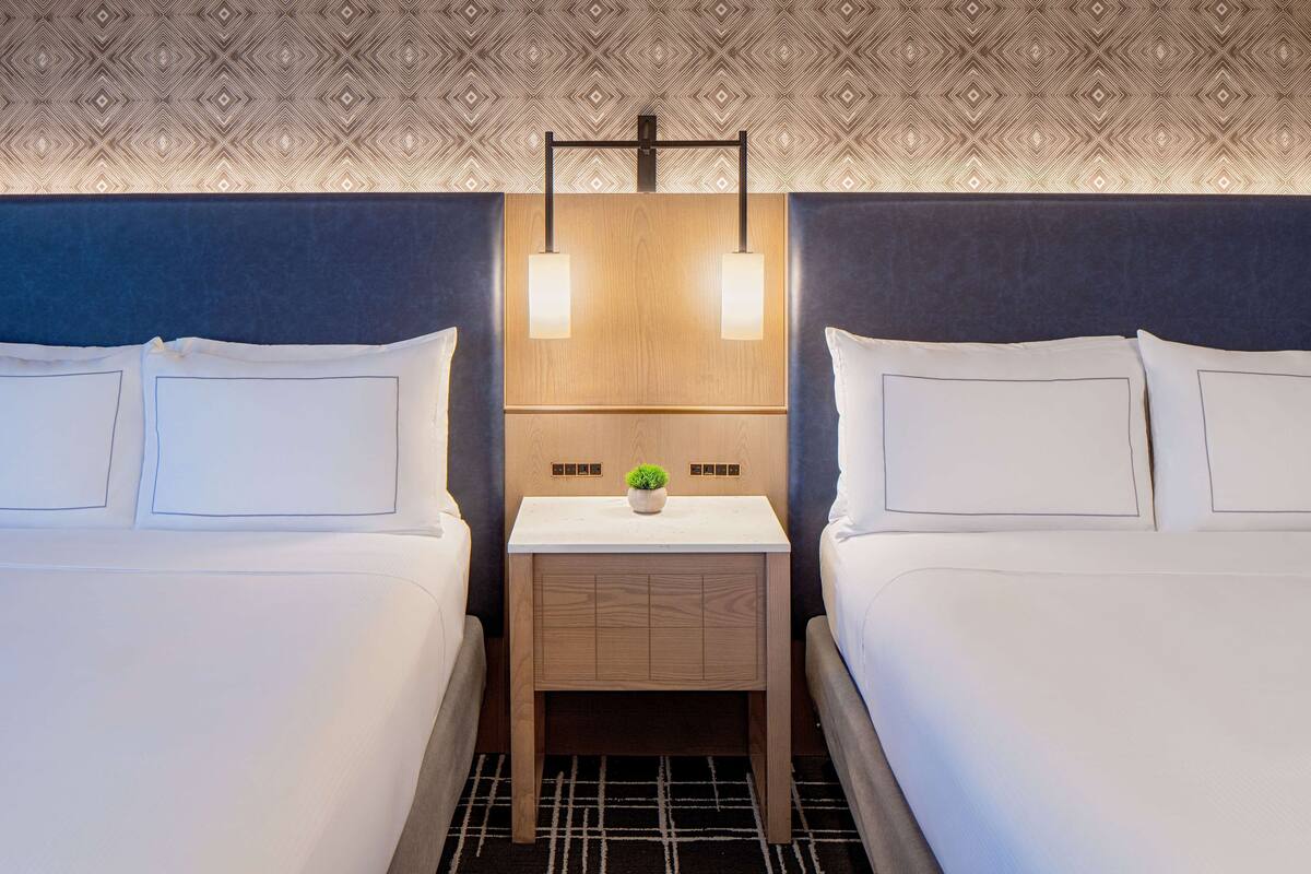A hotel room with two beds, a small wooden nightstand with a plant, and a patterned ceiling.