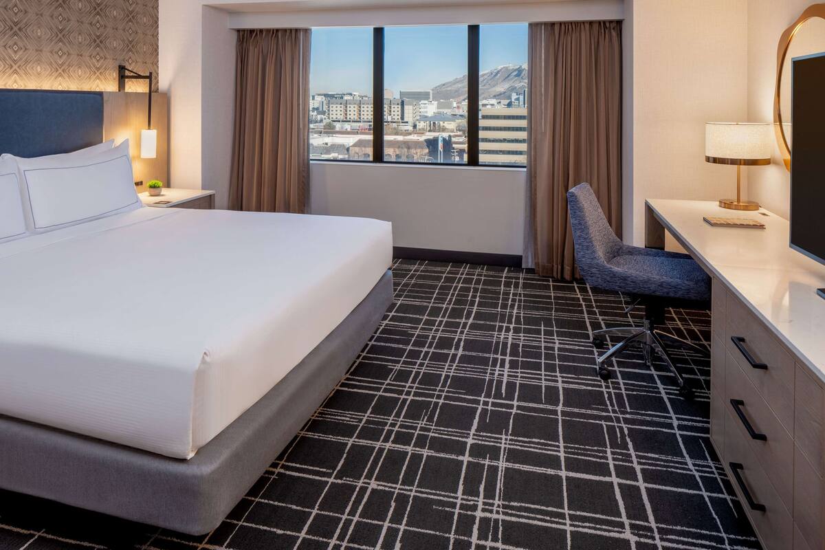 A hotel room with a bed, a desk, a chair, a large window with a city view, and a patterned carpet.