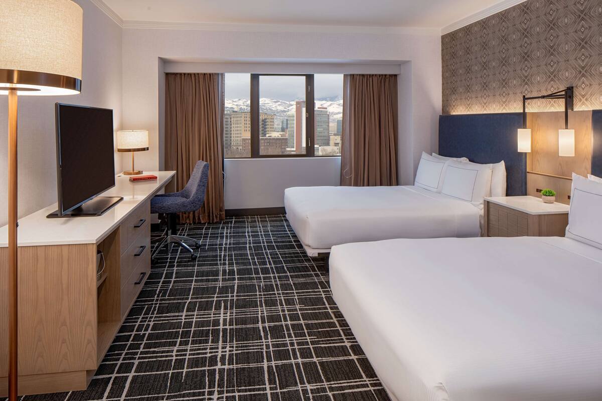 A hotel room with two beds, a desk with a TV, a chair, and a view of a cityscape with snow-capped buildings.