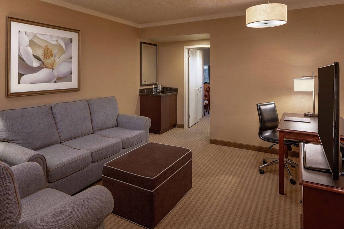 A hotel room with a gray sofa, a wooden coffee table, a desk with a chair, a television, a mirror, and a framed picture on the wall.