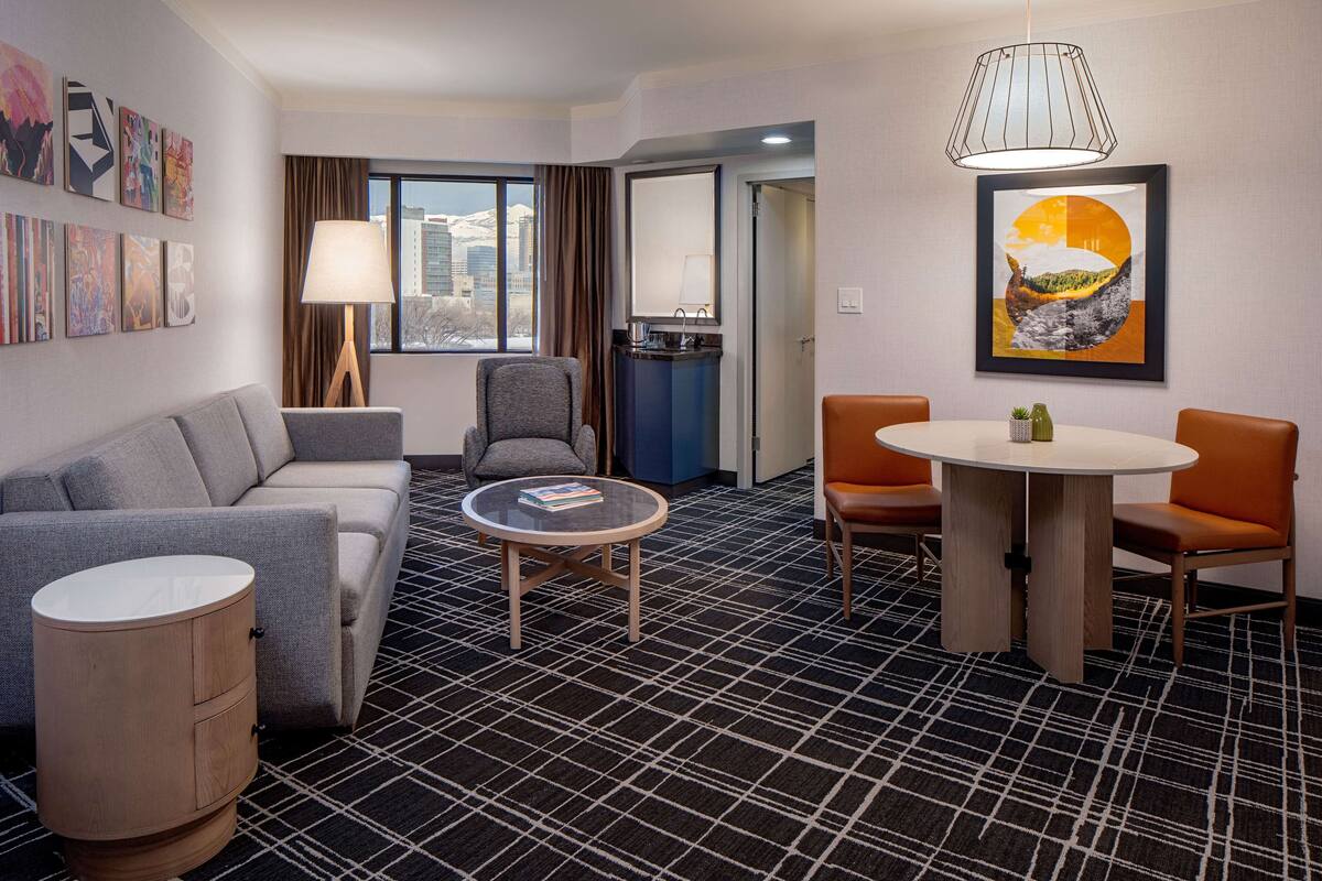 A hotel room with a sofa, armchair, round table, and a cabinet. There is a window with a view of buildings and a painting on the wall.