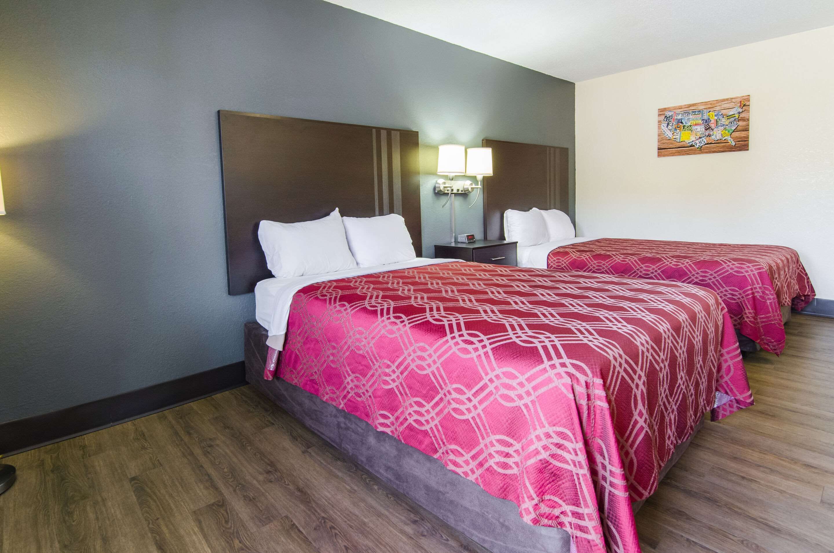 A hotel room with two beds, each with a red patterned bedspread, a nightstand with a lamp, and a wall-mounted map.