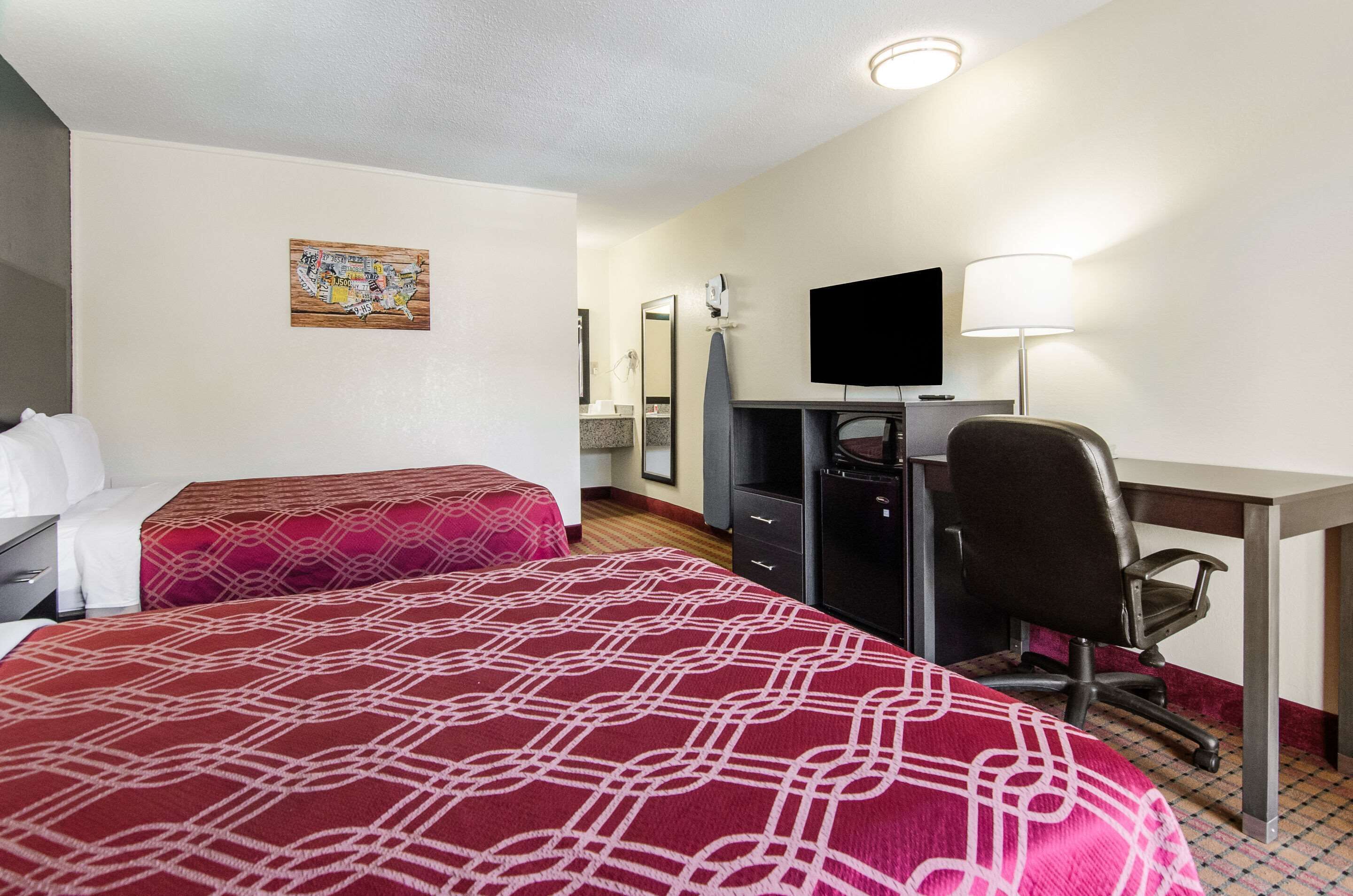 A hotel room with two beds, a desk, a chair, a TV, and a lamp.