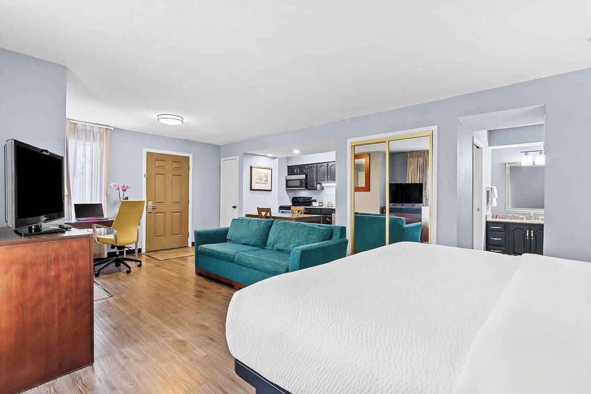 A hotel room with a bed, a desk with a computer, a sofa, a TV, and a kitchenette.