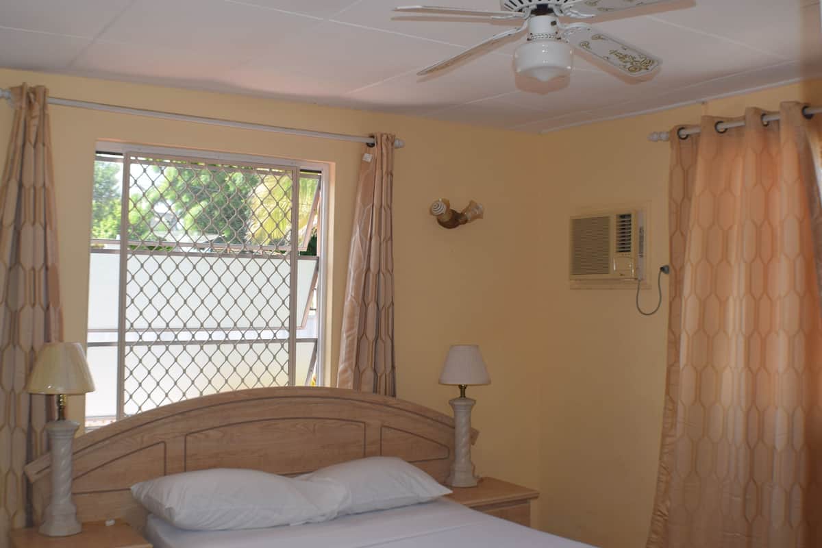 A bedroom with a bed, two bedside lamps, a ceiling fan, a window with curtains, and a wall-mounted air conditioning unit.