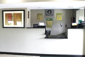 Mostrador de recepción de hotel con una obra de arte enmarcada, un cartel de 'Quality by Choice Hotels', una tarjeta de fidelización 'Choice' y un quiosco digital, además de un teléfono.