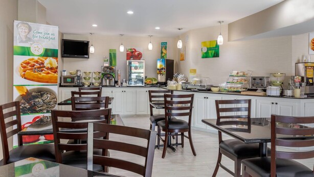A modern breakfast area with wooden chairs and tables, a flat-screen TV, and a variety of breakfast items displayed on the counter.