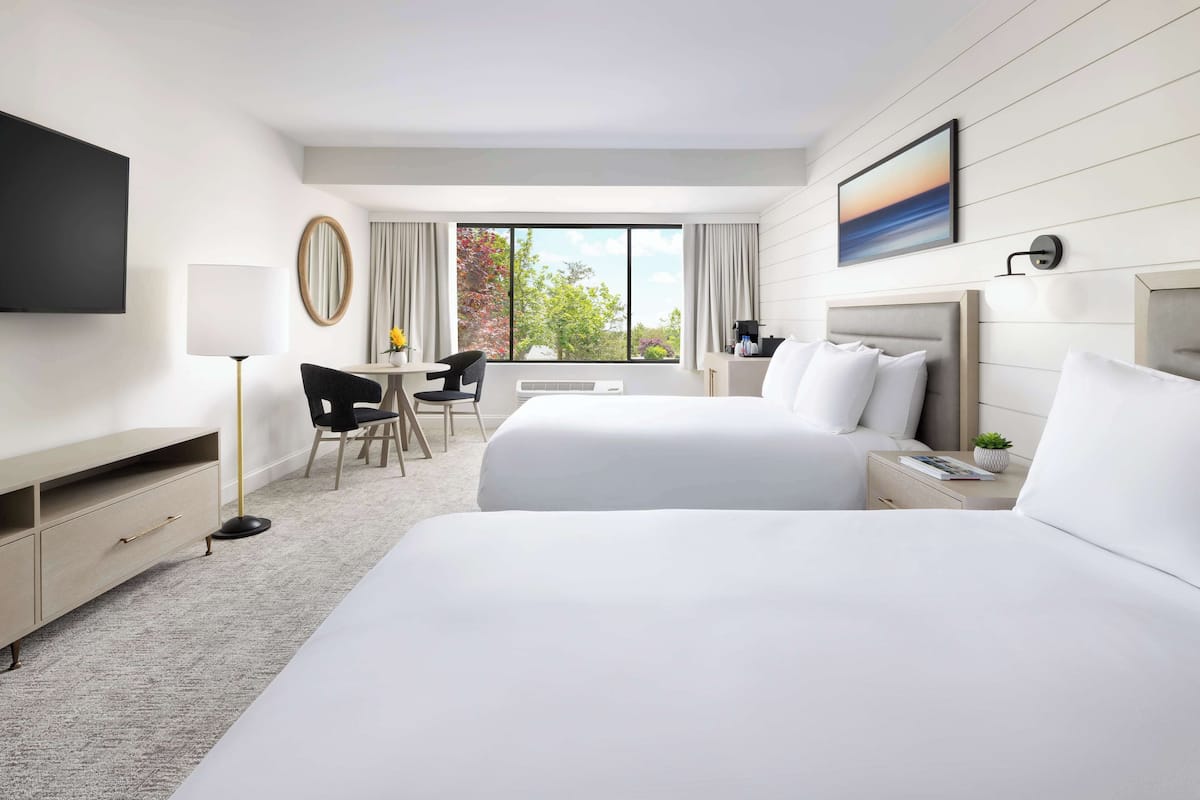 A hotel room with two beds, a desk, a chair, a TV, and a large window with curtains.