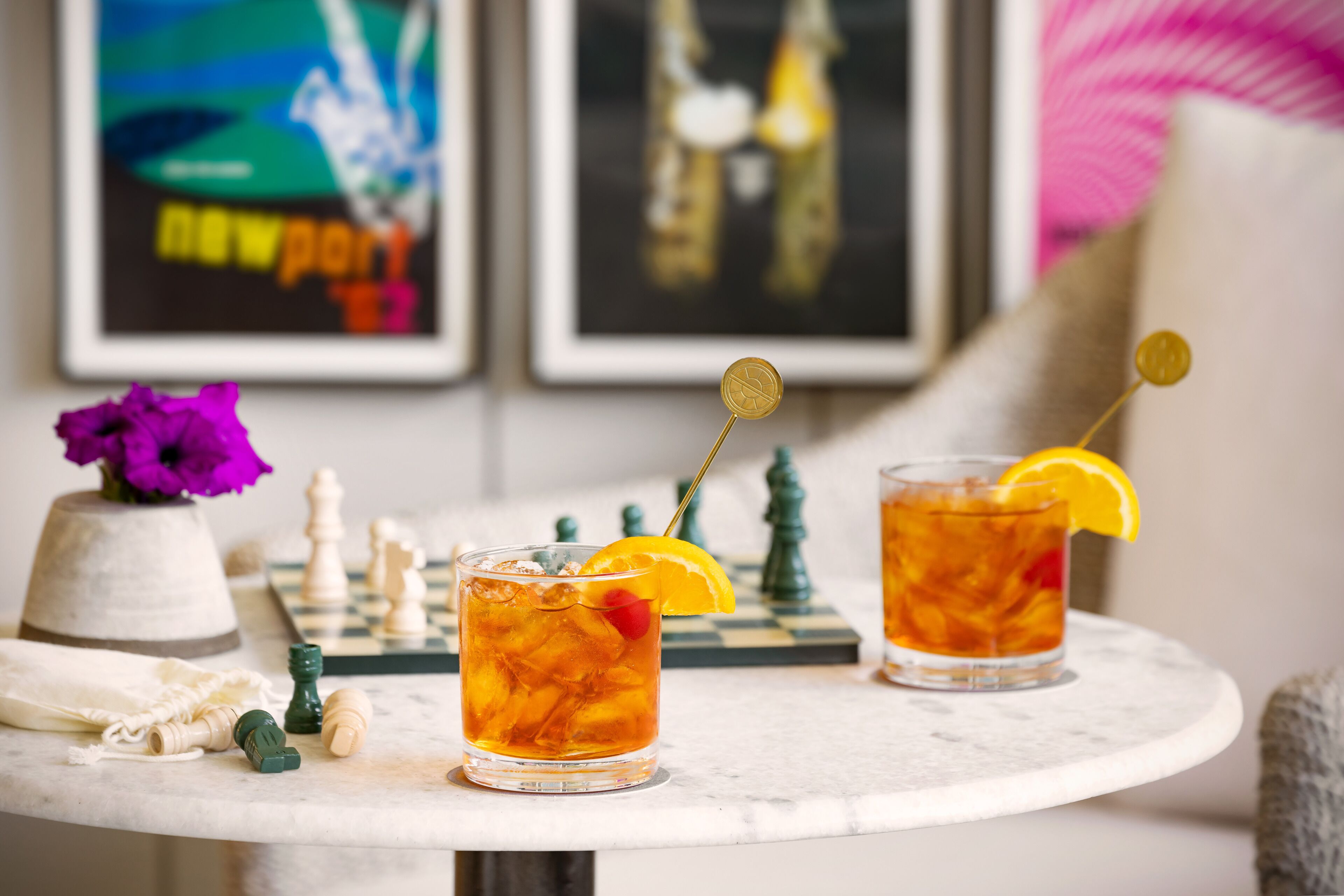 Dos vasos de té helado con rodajas de naranja sobre una mesa de mármol, piezas de ajedrez y un florero con flores moradas.