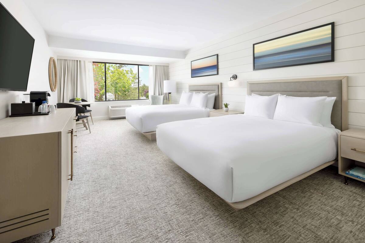 A hotel room with two beds, a desk, a chair, a TV, and a large window with a view of trees.