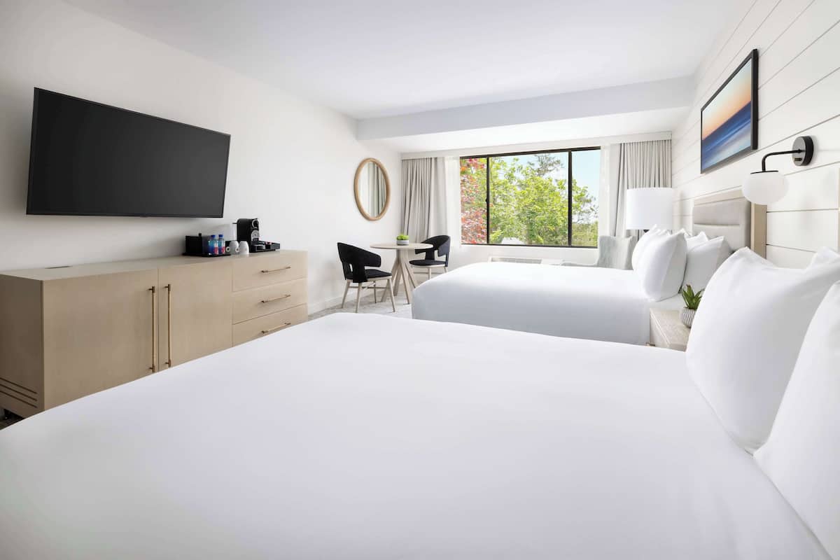 A hotel room with two beds, a TV, a desk with chairs, a window with a view, and a flat-screen TV mounted on the wall.