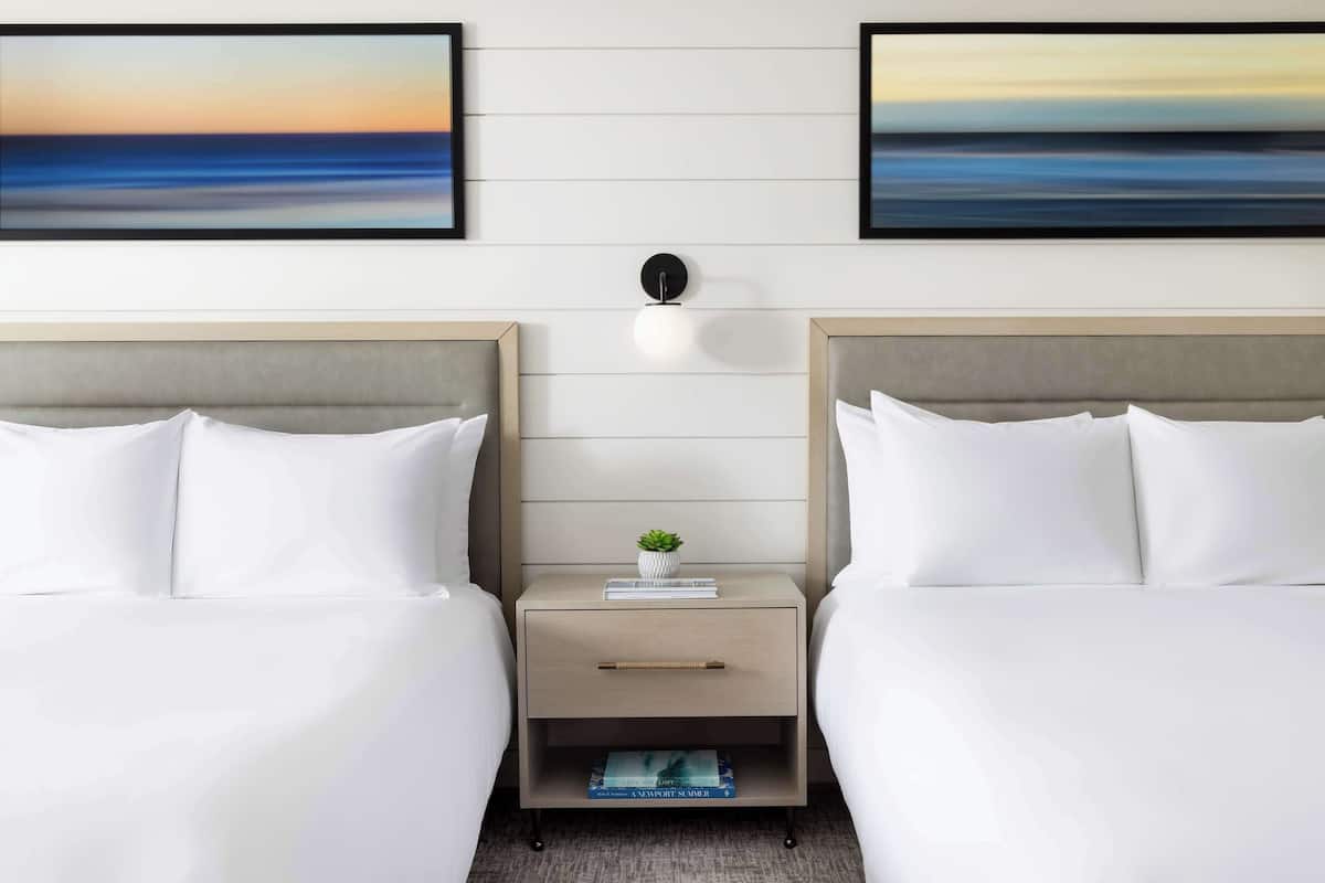 A hotel room with two beds, a nightstand with a lamp, and a small shelf with books.