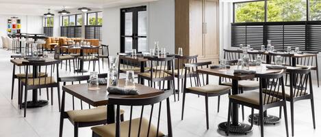 Un área de comedor moderna con varias mesas y sillas, ventanas grandes y un armario de madera.