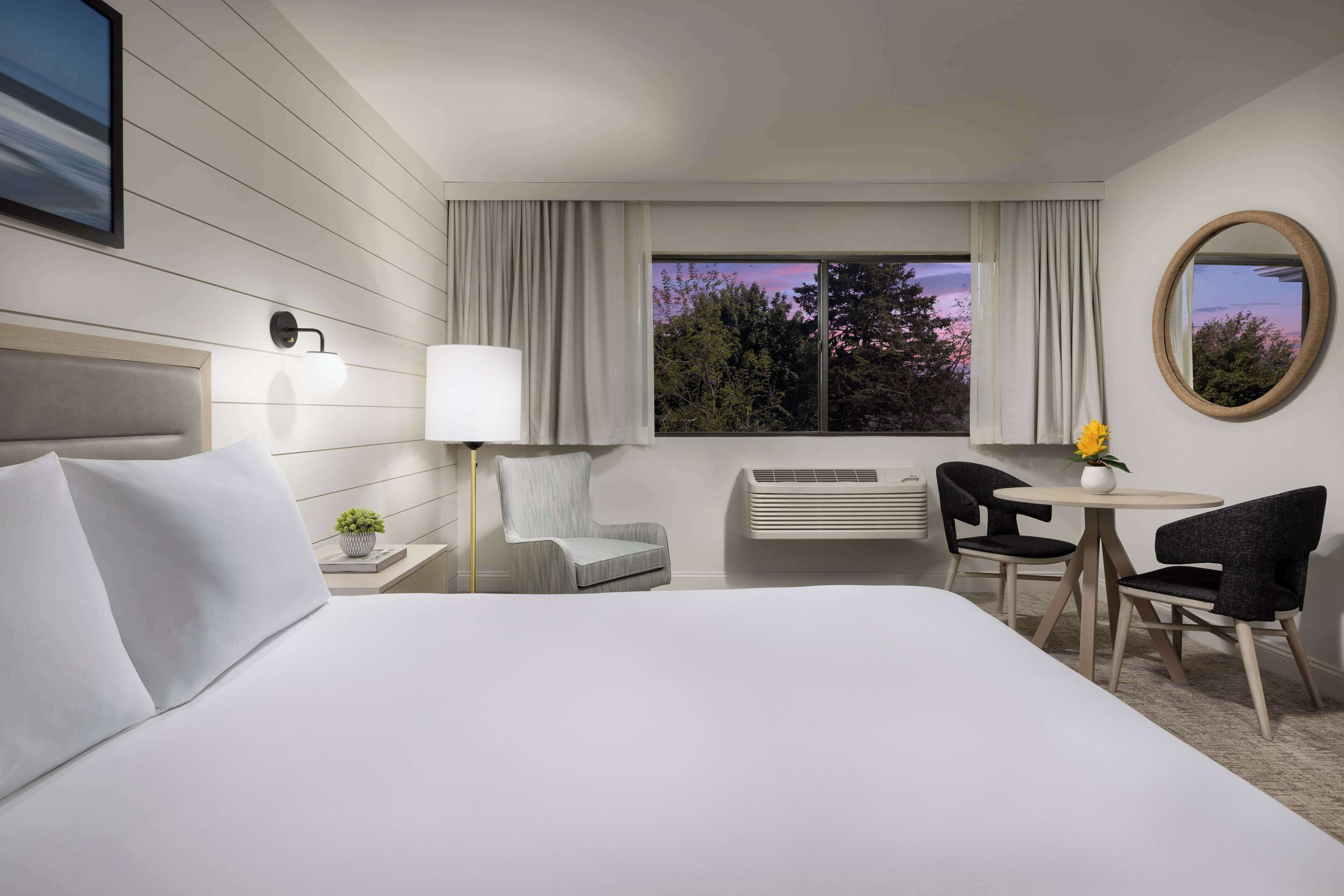 A hotel room with a large bed, a desk with two chairs, a window with curtains, and a wall-mounted mirror.