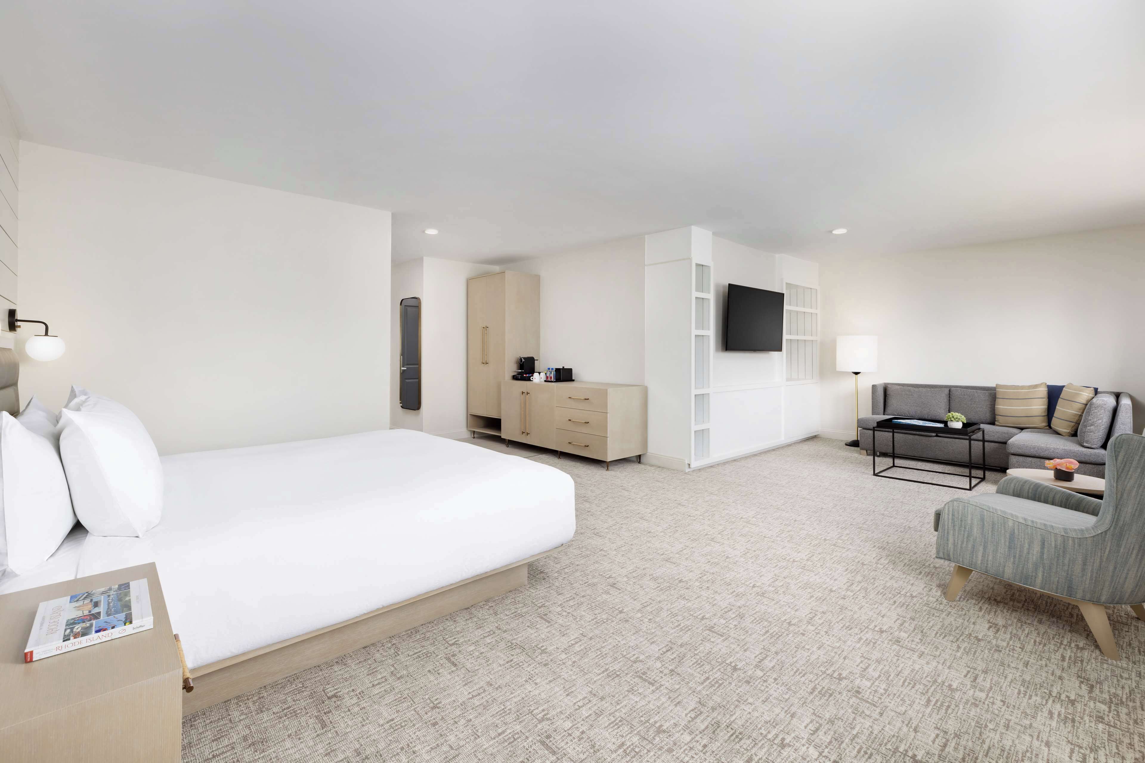 A modern hotel room with a large bed, a sofa, a TV mounted on the wall, and a small table with a lamp.