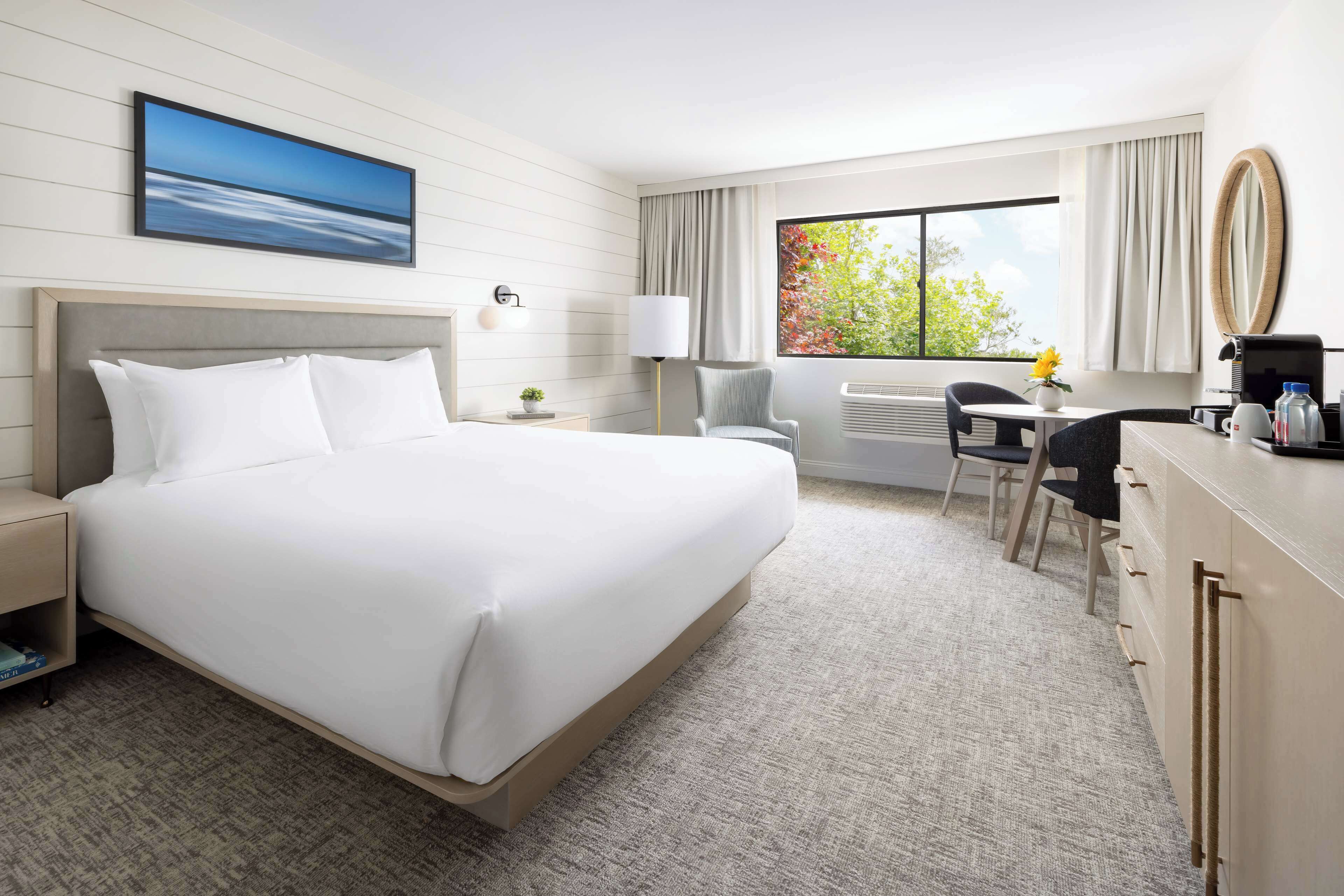 Habitación de hotel con una cama grande, un escritorio con silla, una mesita con un florero, una lámpara y una ventana con cortinas.