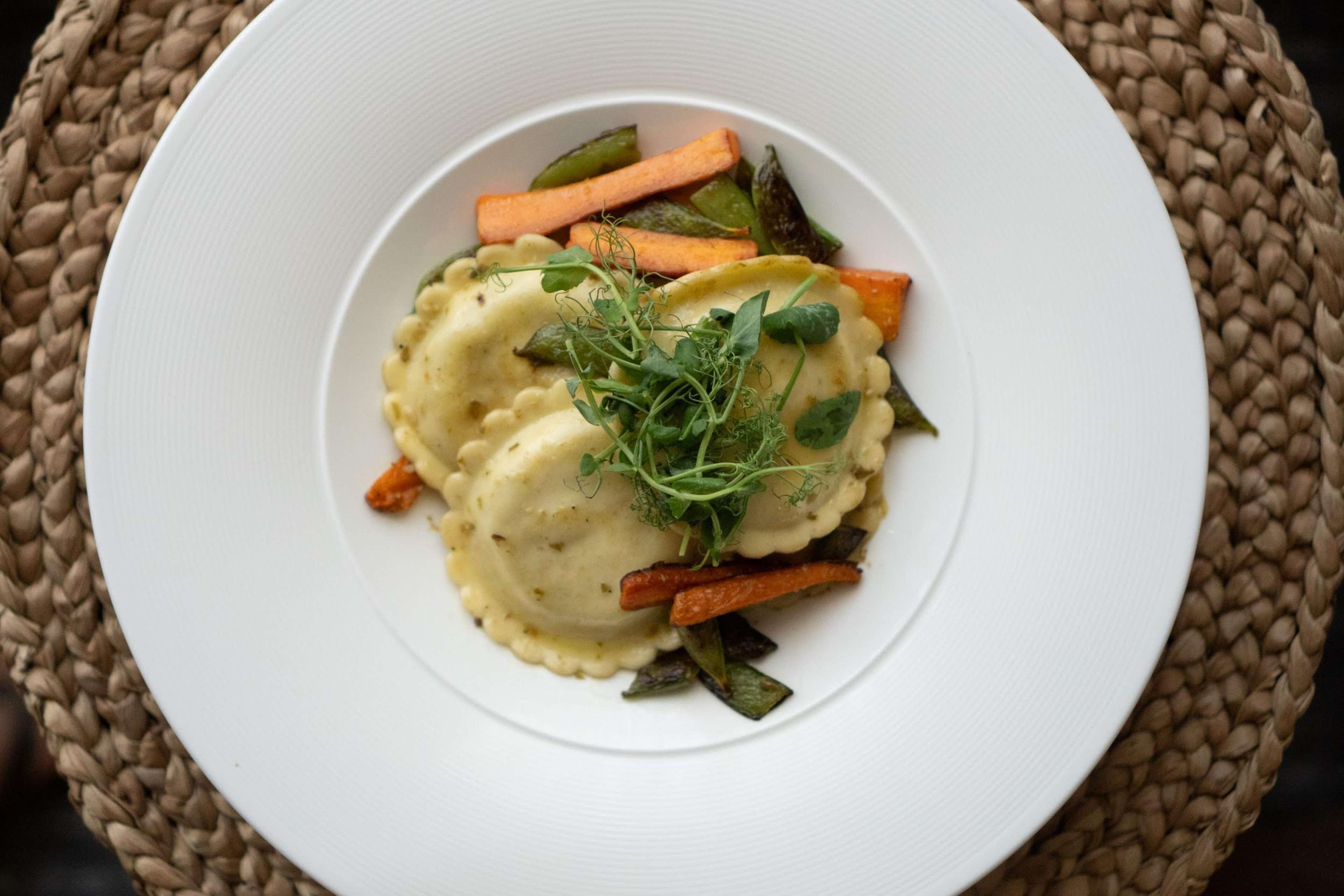 Une assiette blanche contenant des raviolis, des carottes et des légumes verts, le tout posé sur un set de table en fibres tressées.
