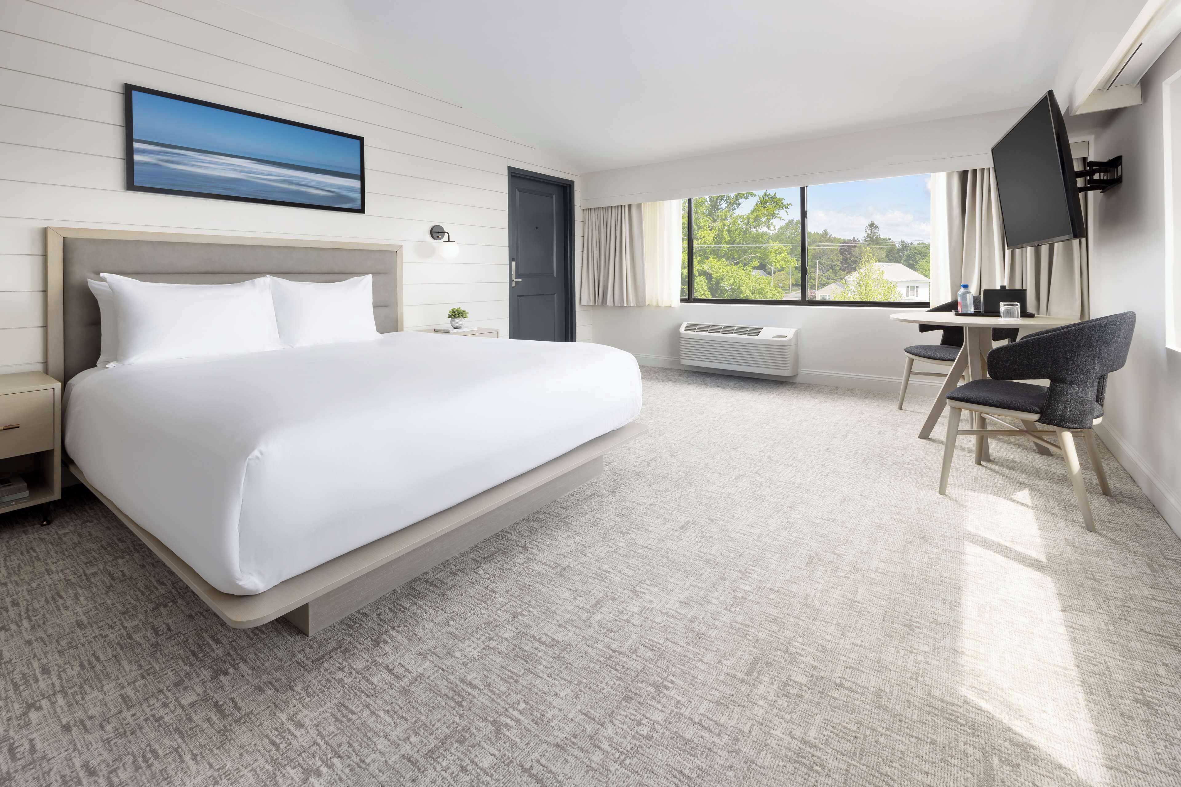 A hotel room with a large bed, a desk with a chair, a TV, and a window with a view of greenery.