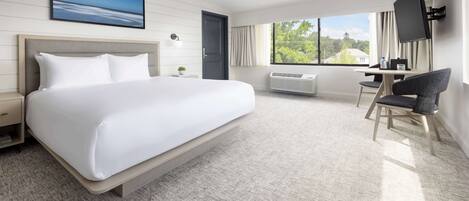A hotel room with a large bed, a desk with a chair, a TV, and a window with a view of greenery.