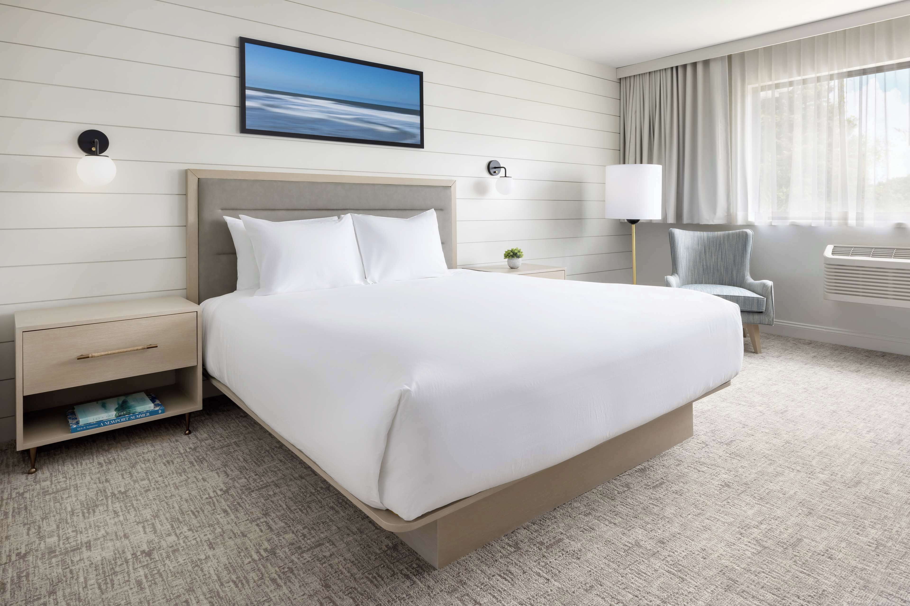 A hotel room with a large bed, a nightstand, a chair, a lamp, and a window with curtains.