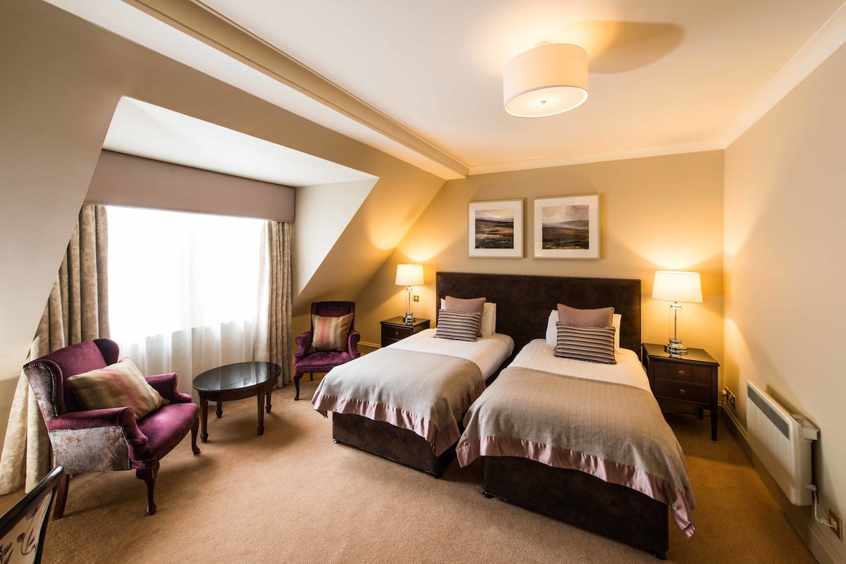A hotel room with two beds, a chair, a small table, and a lamp.