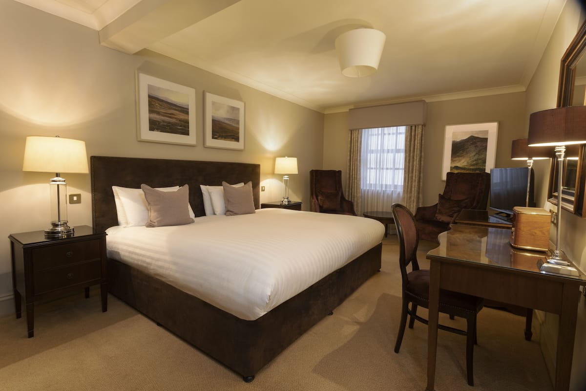 A hotel room with a large bed, two bedside tables with lamps, a desk with a chair, and two framed pictures on the wall.