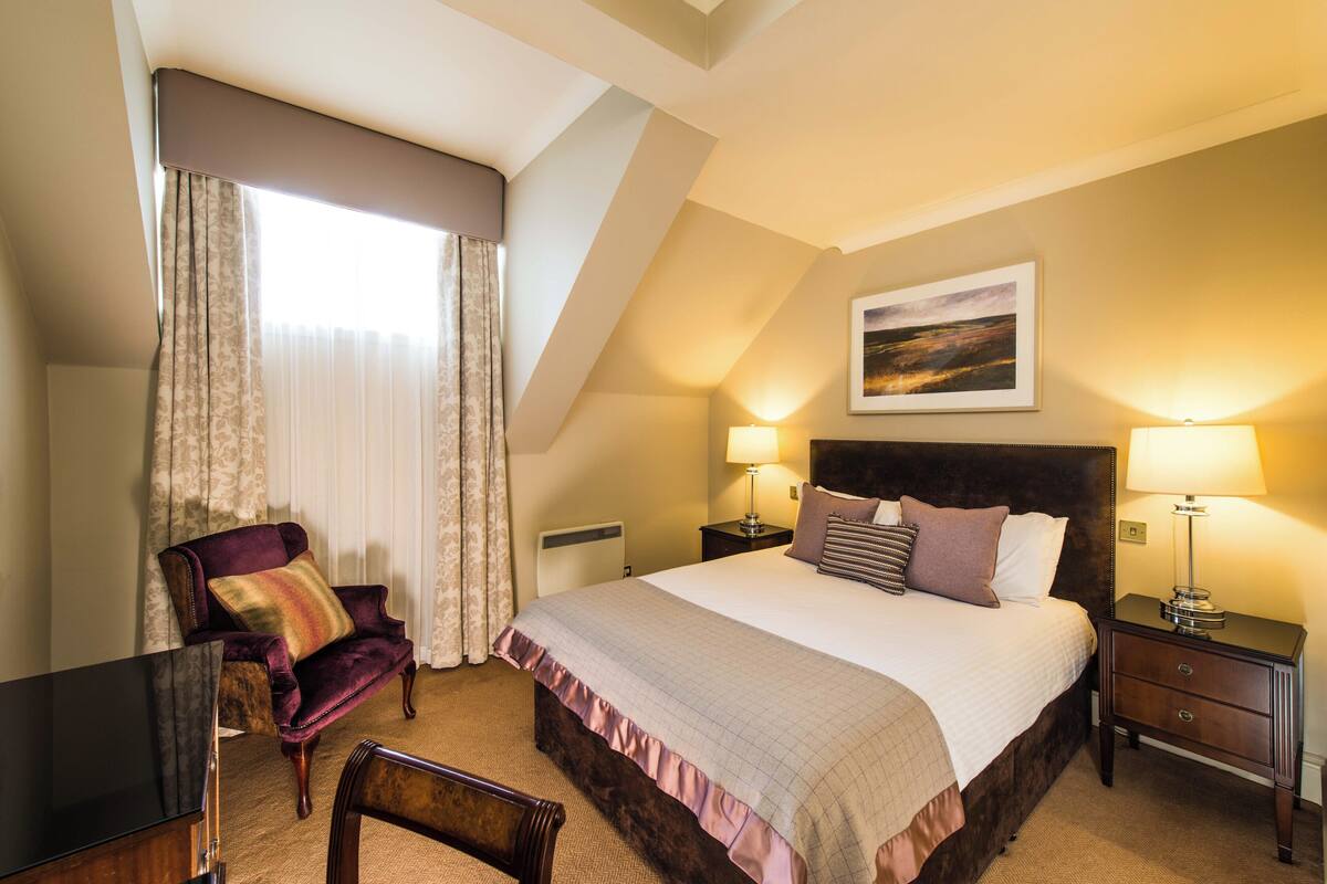 A hotel room with a bed, two bedside tables with lamps, a chair, a desk, and a painting on the wall.