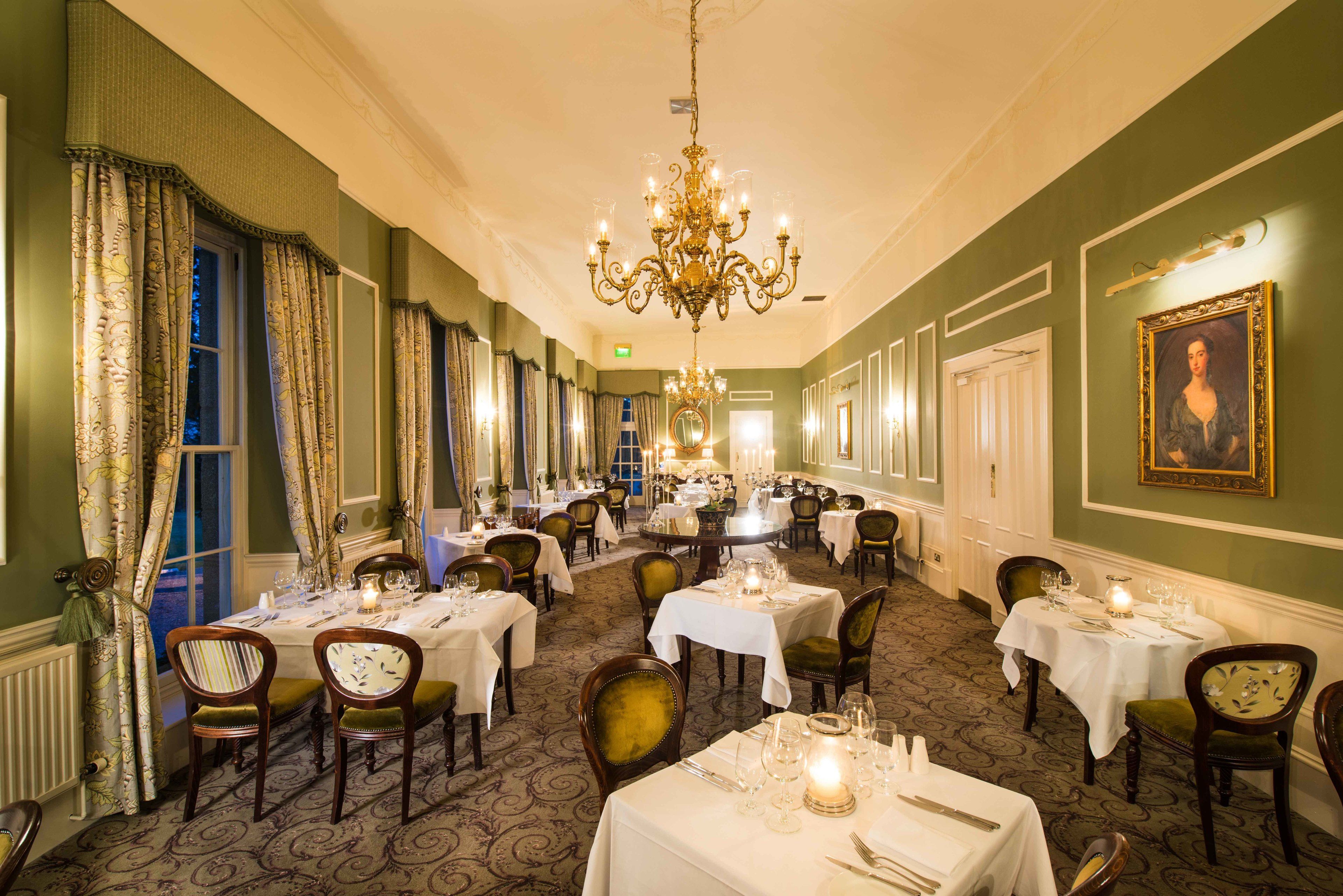 A formal dining room with multiple tables set for service, a chandelier, and a portrait on the wall.