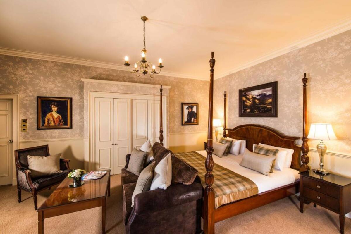 A four-poster bed with a wooden headboard, a sitting area with a sofa and armchair, a wooden coffee table with a vase, and a nightstand with a lamp.