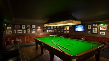 Une table de billard dans une pièce à la lumière tamisée, avec des tableaux encadrés aux murs, un téléviseur et un coin bar.