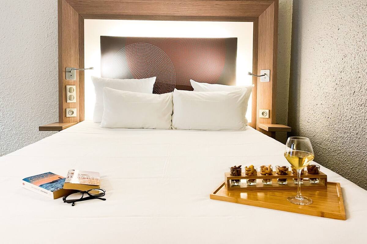 A neatly made bed with white linens, a bedside table with a tray of snacks and a glass of wine, and a book and glasses on the bed.