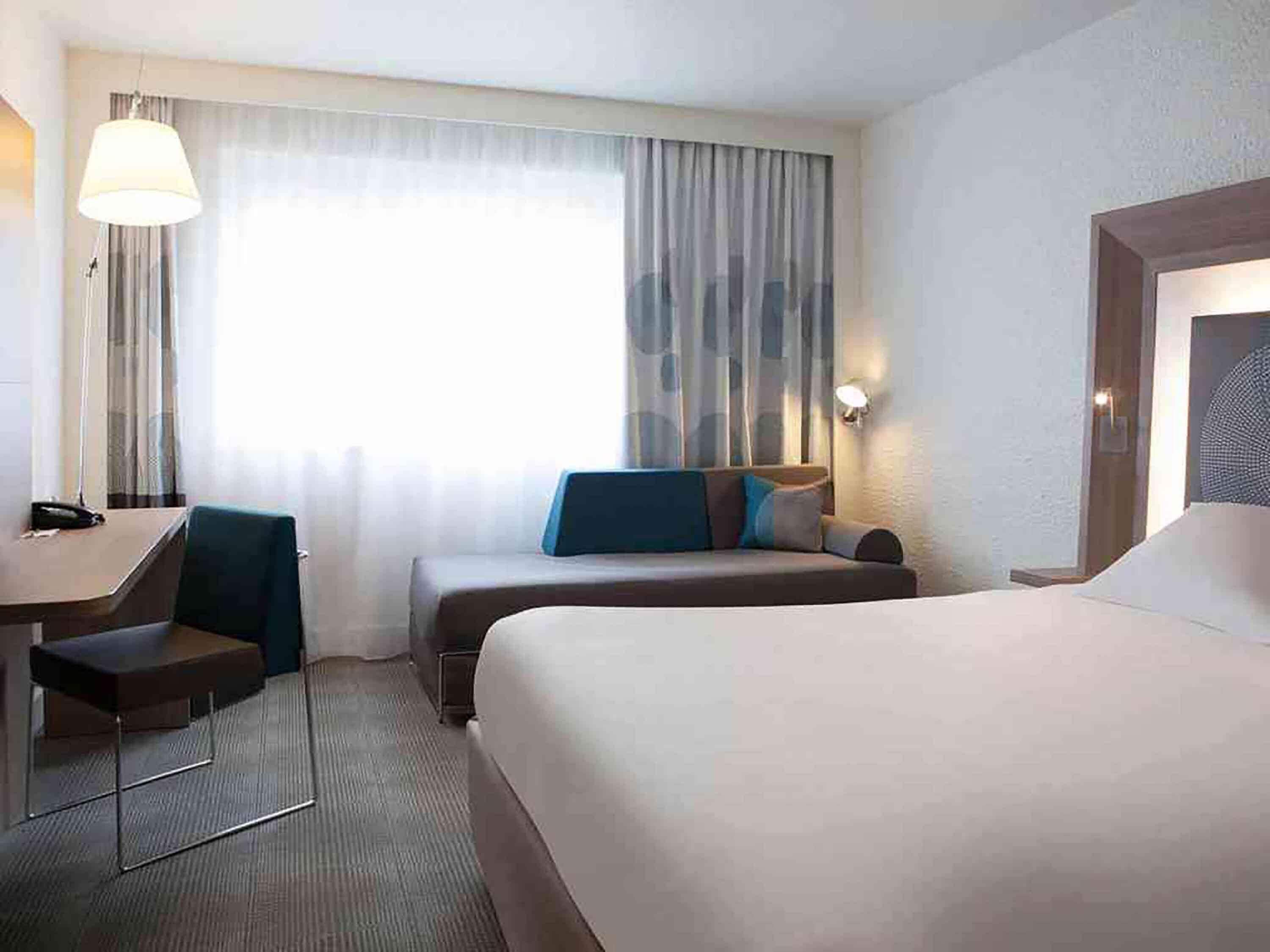A hotel room with a bed, a desk, a chair, a lamp, a window with curtains, and a wall-mounted light.