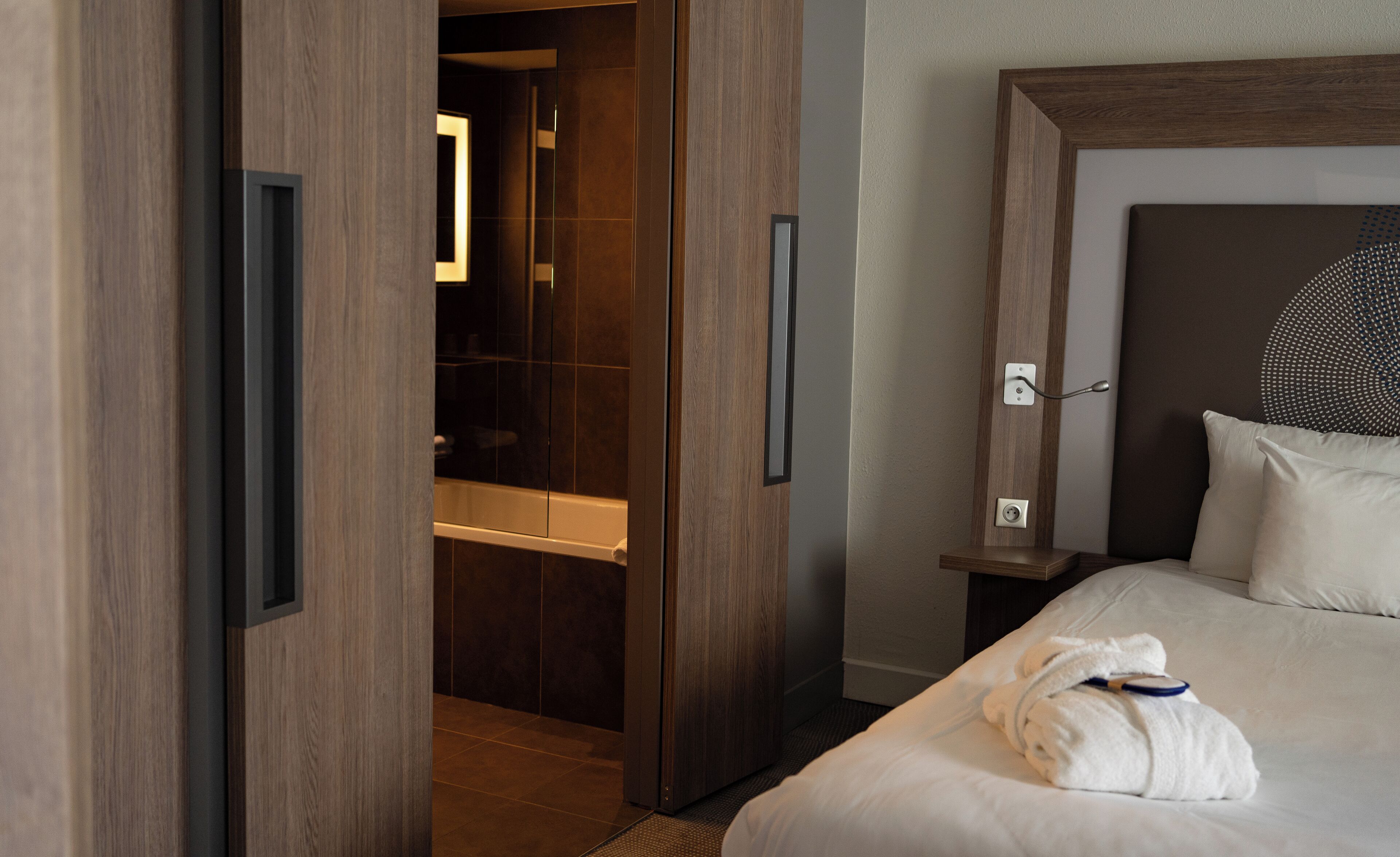 A hotel room with a wooden door, a shower area, a bedside table with a lamp, and a neatly made bed with a towel.