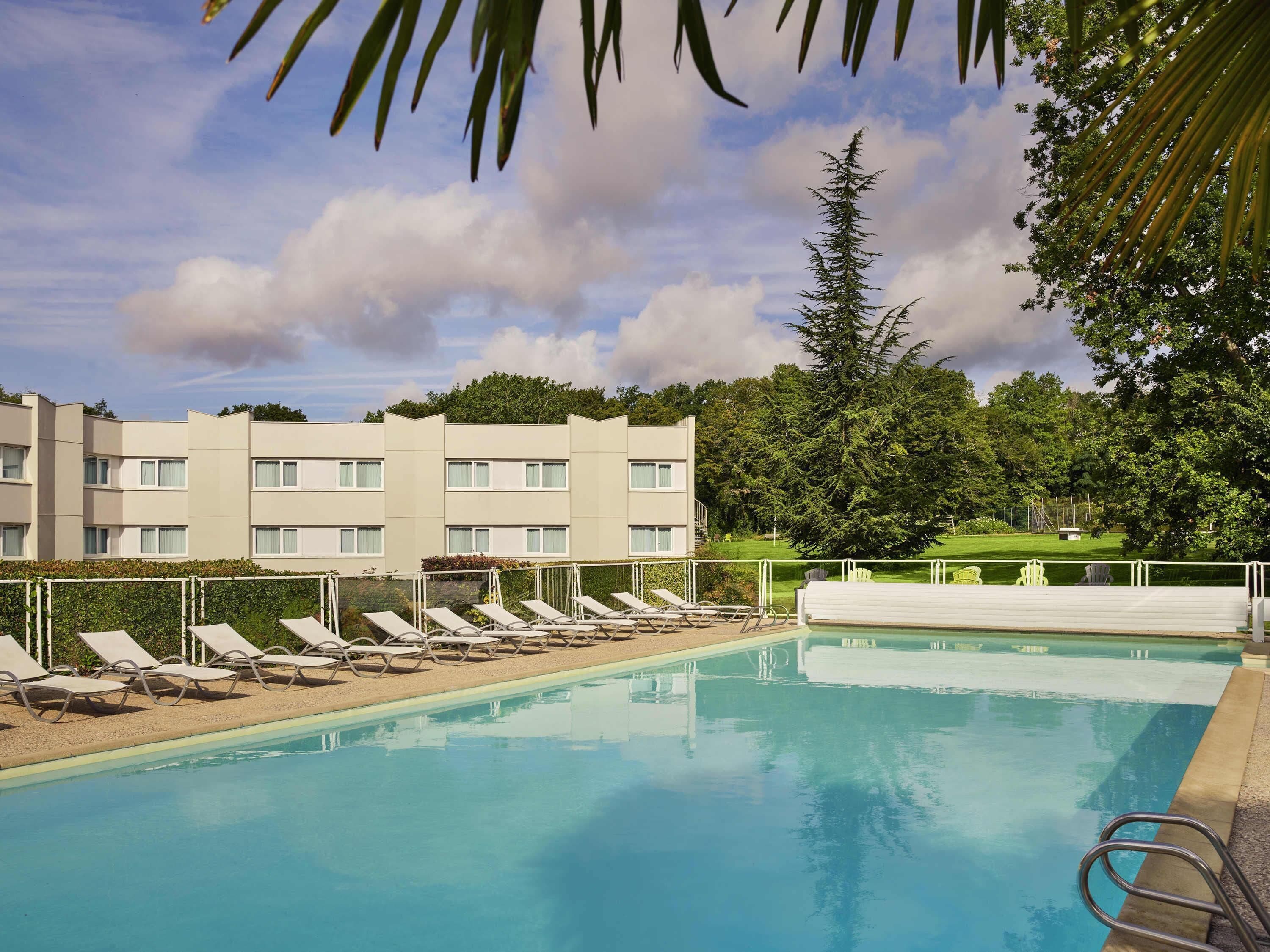 A swimming pool with lounge chairs, a building with multiple windows, and a tree in the background.