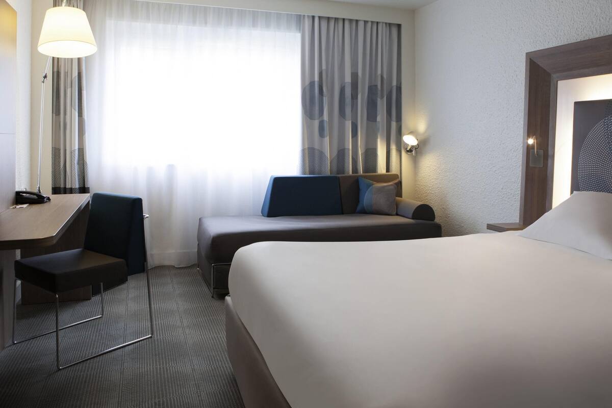 A hotel room with a bed, a desk, a chair, a sofa, and a window with curtains.