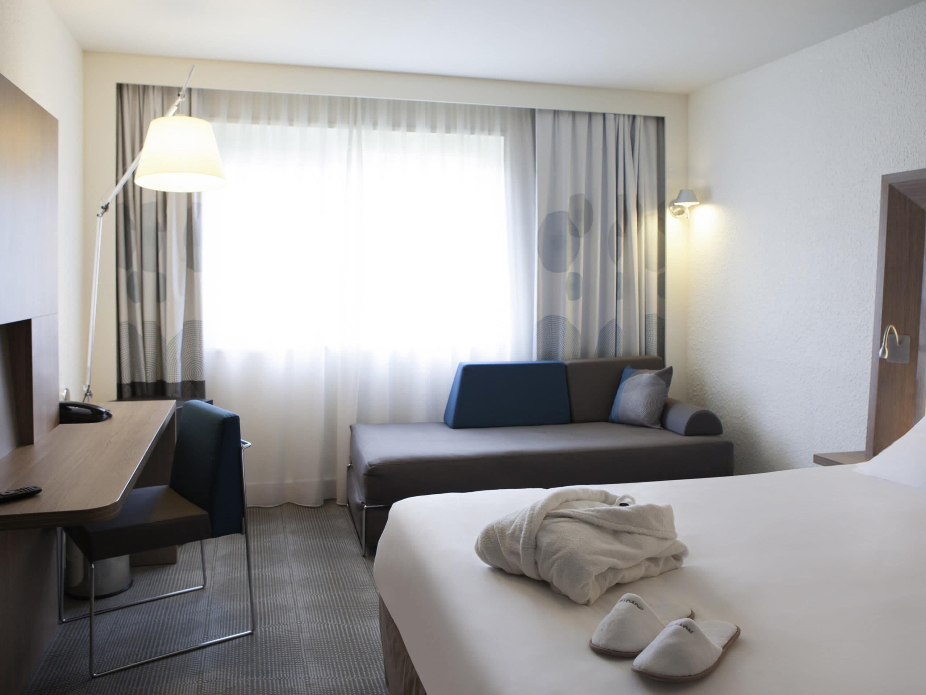 Une chambre d’hôtel comprenant un lit, un bureau, une chaise, un canapé, une lampe et une fenêtre avec des rideaux.
