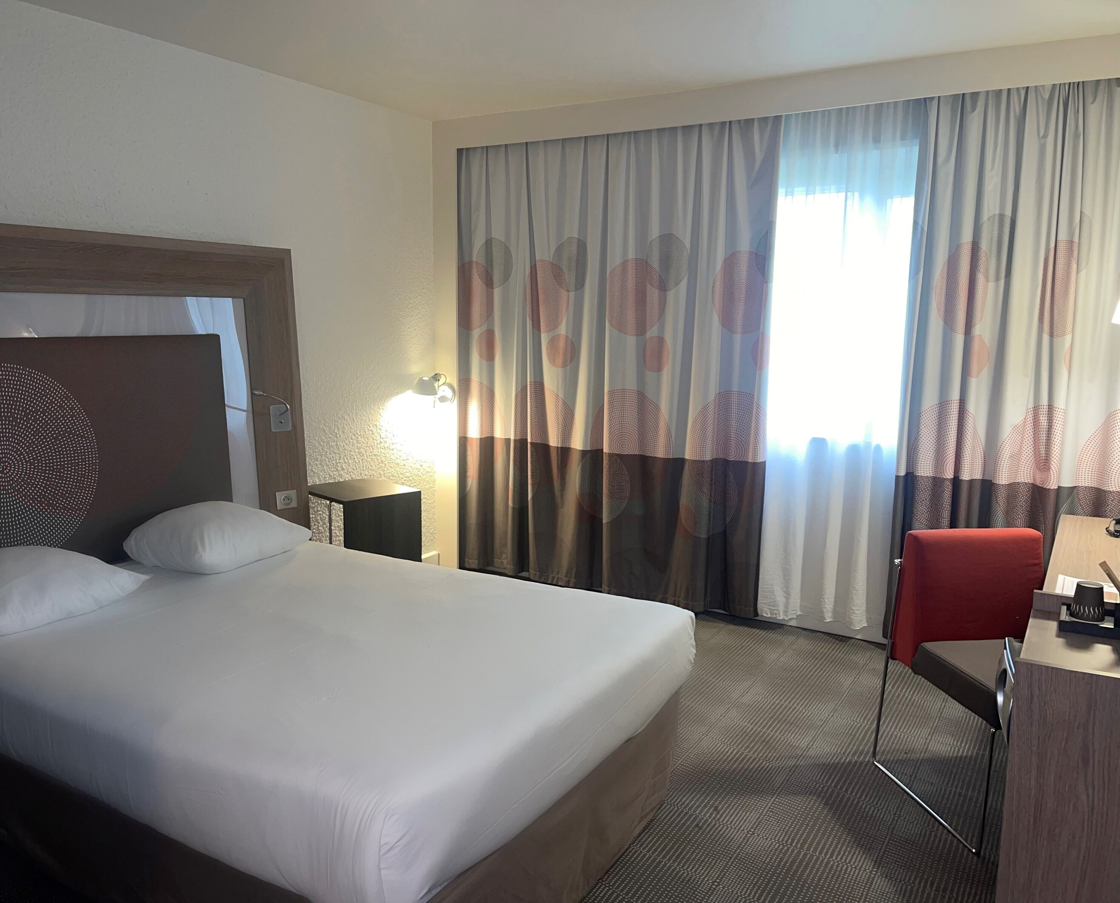 A hotel room with a large bed, a desk, a chair, a lamp, and a window with curtains.